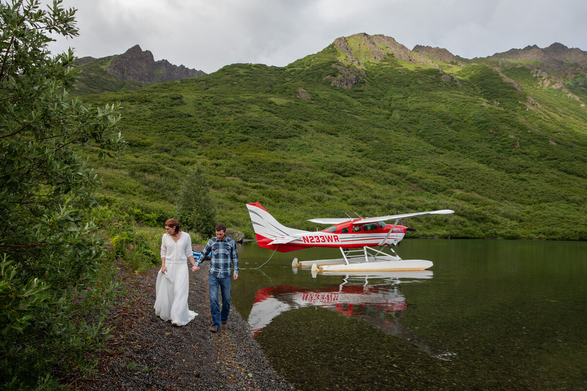 A bride and groom walk along a rocky beach hand in hand as a float plane sits on the lake behind them.