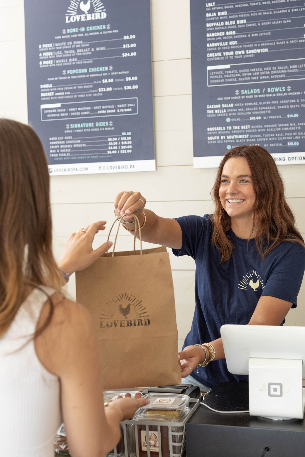 young woman behind counter handing bag of food to customer with menus on the wall behind her