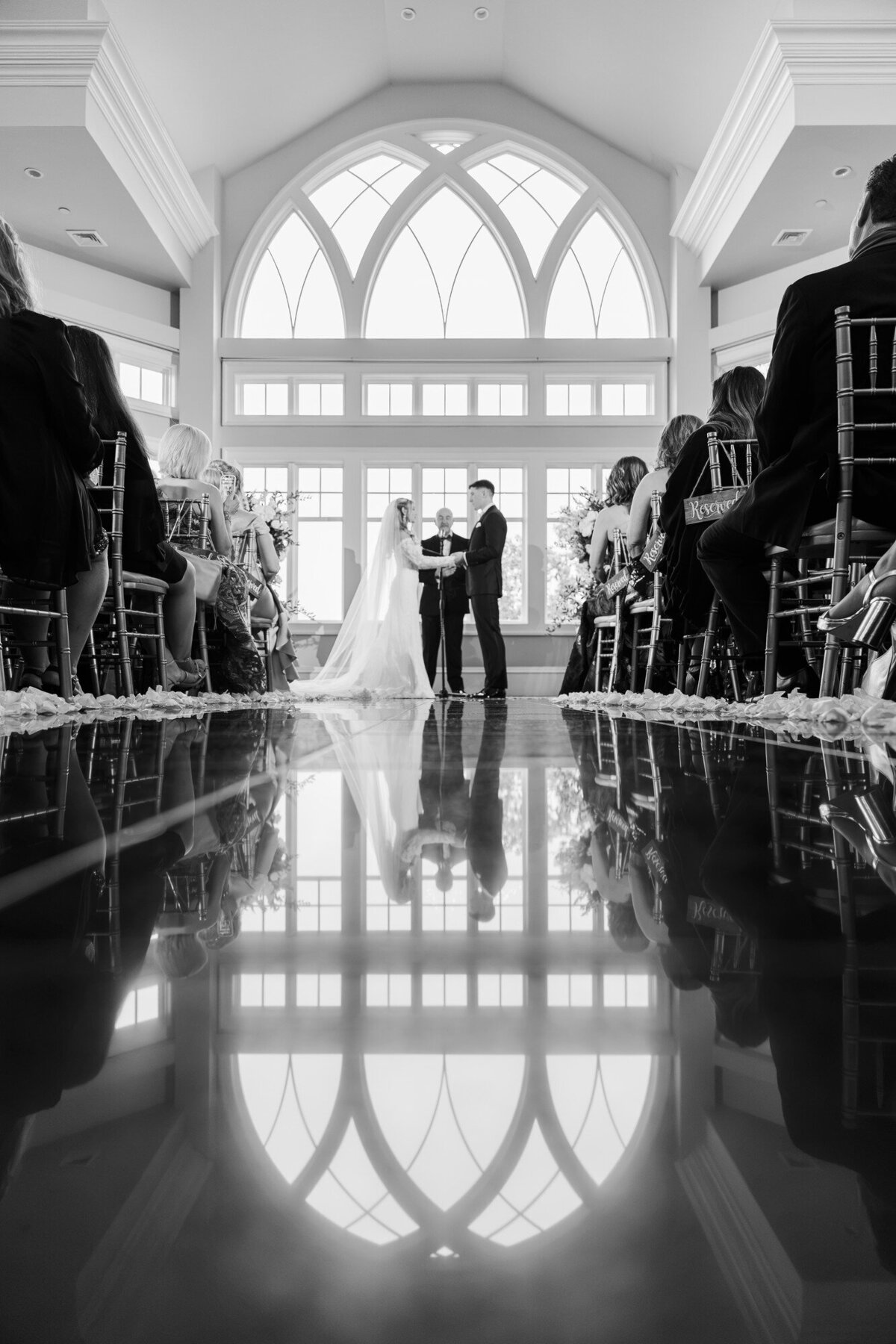 Black and white wide shot of a couple at the end of a church aisle with arched windows reflected on the floor