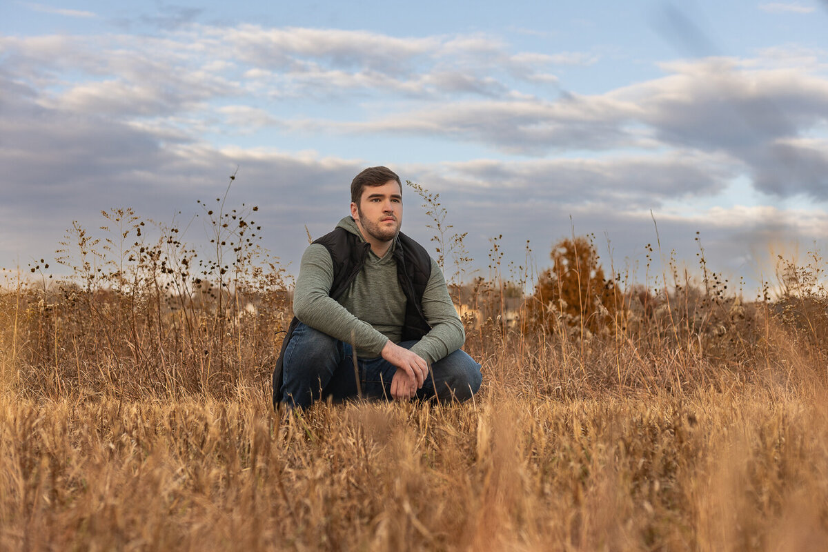 A senior guy squatting in a grass field looking off in the distance in Lawrence, KS