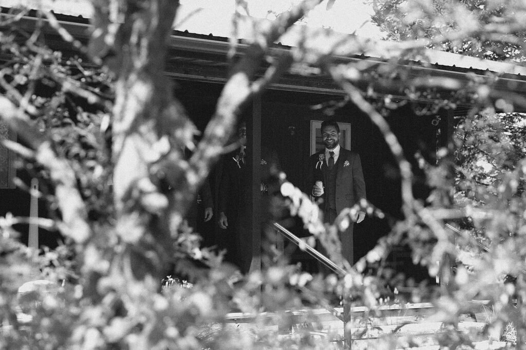 black and white image of a groom on the porch of the Willowbrook wedding venue preparation cottage through the lush trees on the property