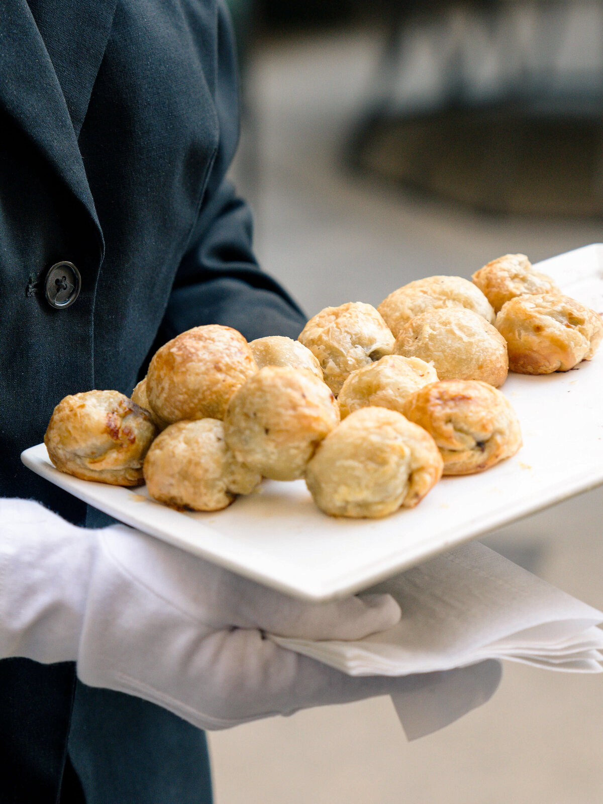 A person in a dark suit and white gloves holds a rectangular white platter with several golden-brown puff pastry hors doeuvres. The background is blurred.