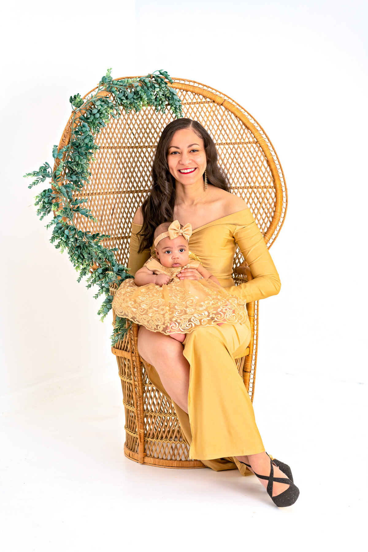 A mom in a gold, formal dress is holding her young baby as they sit in a peacock chair at a photography studio.