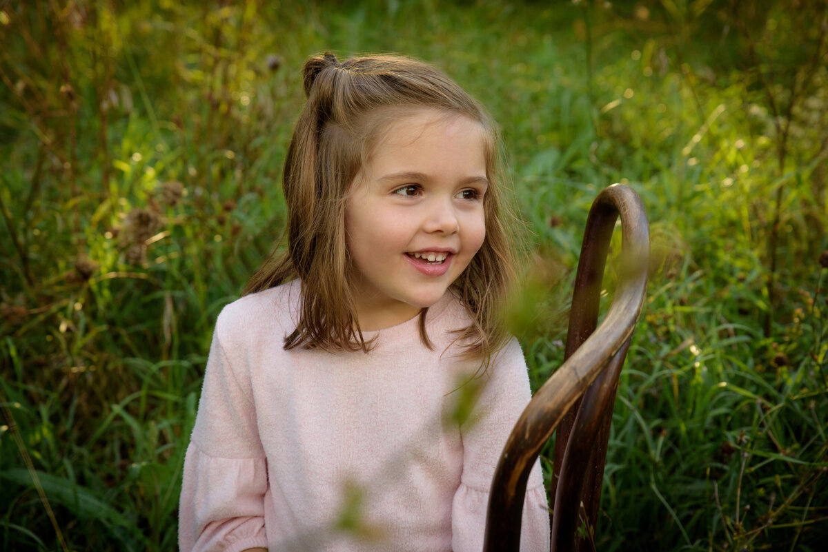 Young girl wearing a pink shirt and jeans sitting in a wooden chair in a long grassy field at Fonferek Glen County Park near Green Bay, Wisconsin