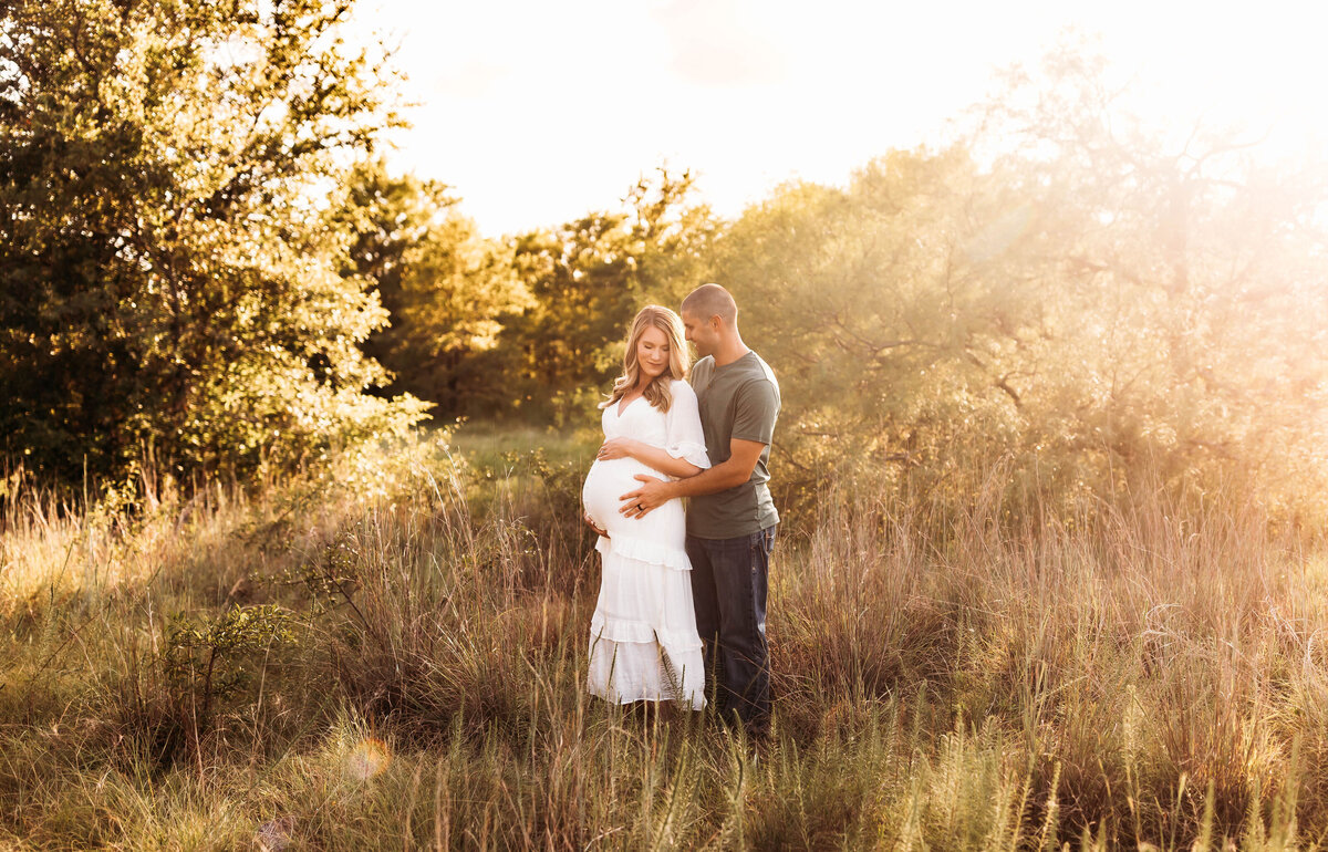 Pregnant couple stands in a field embracing at golden hour.