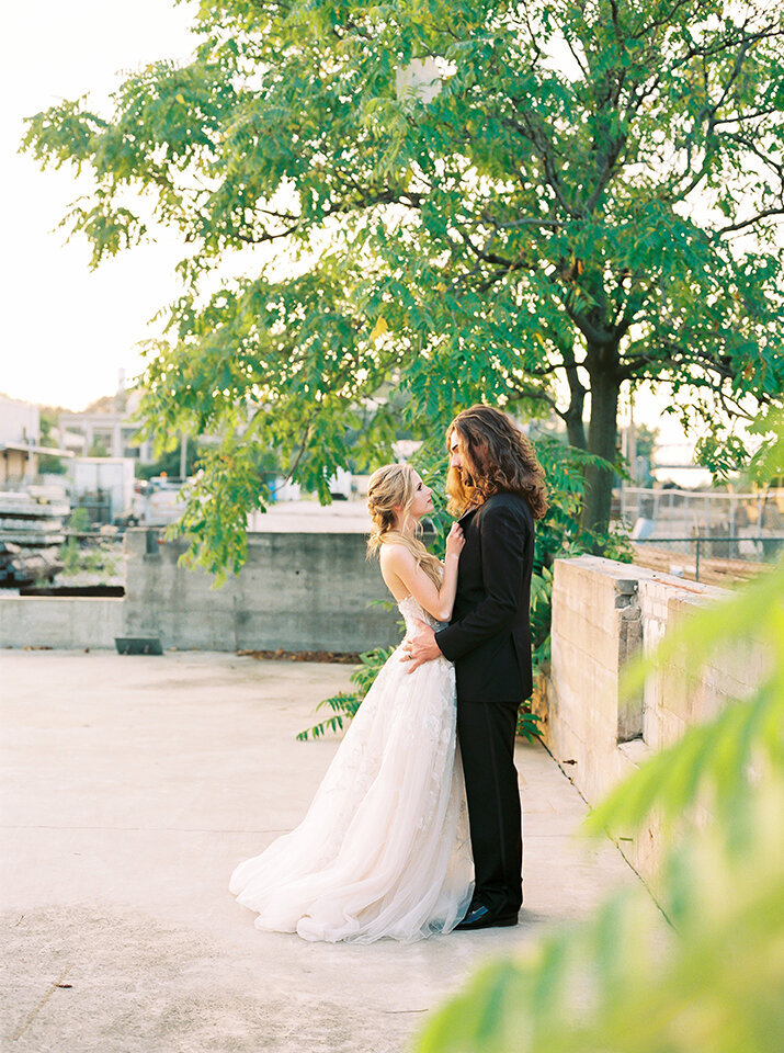 Bride and groom wearing a black tuxedo and white wedding gown holding each other outside on a rooftop.
