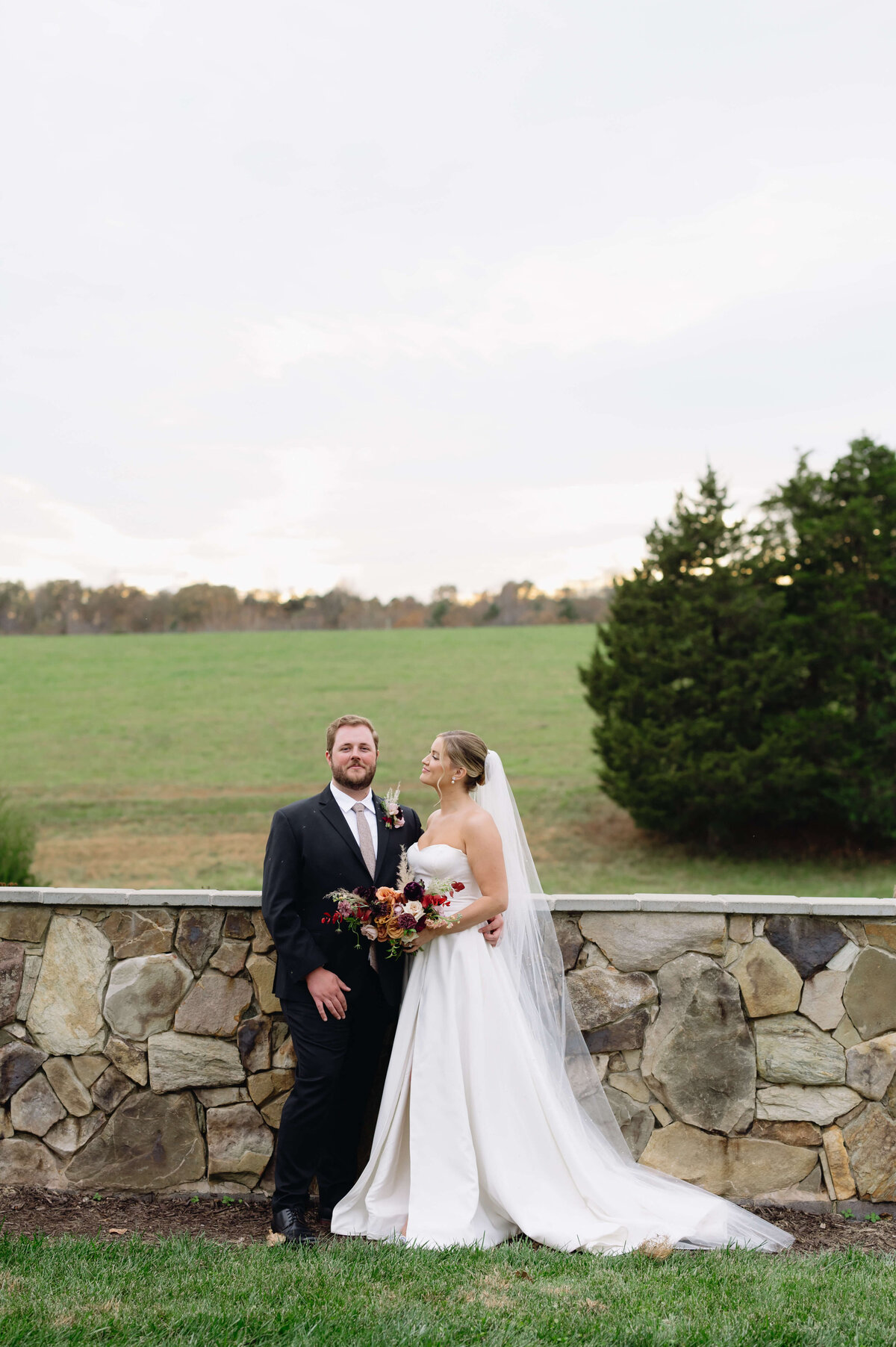 LayZ S Ranch wedding captured by Virginia wedding photographer with bride and groom standing close to each other in front of a stone wall with green hills behind them