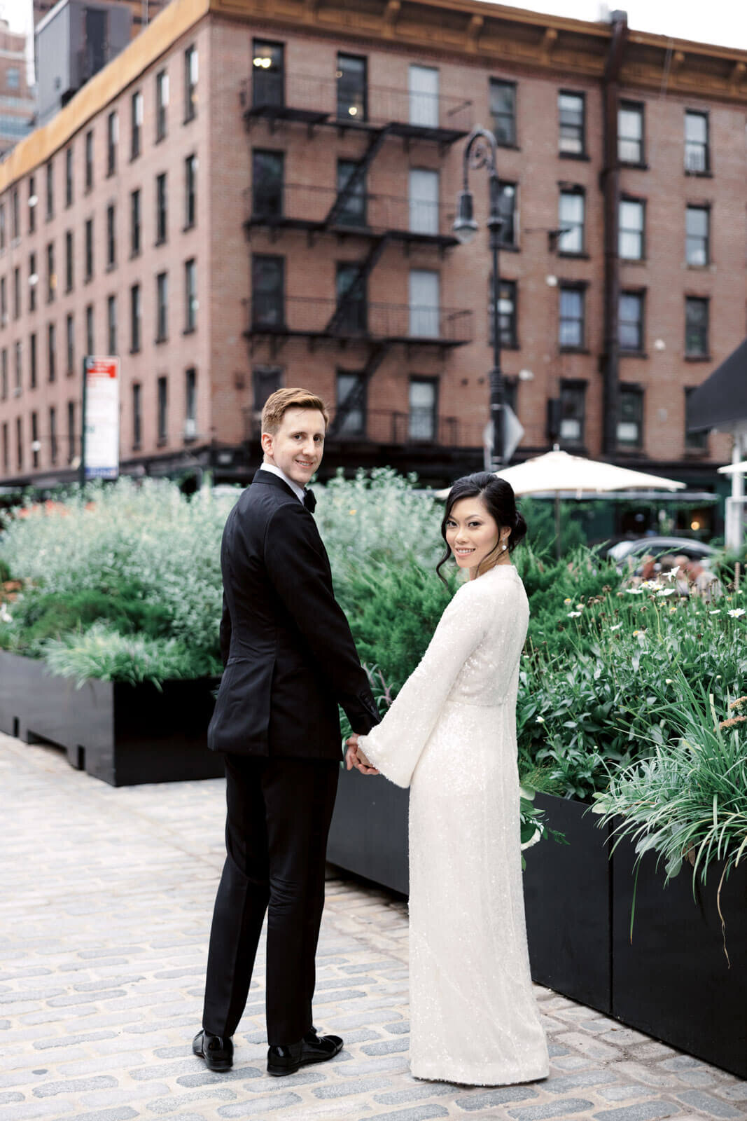 The bride and groom are holding hands, with some plants and a red brick building in the background in West Village, NYC.