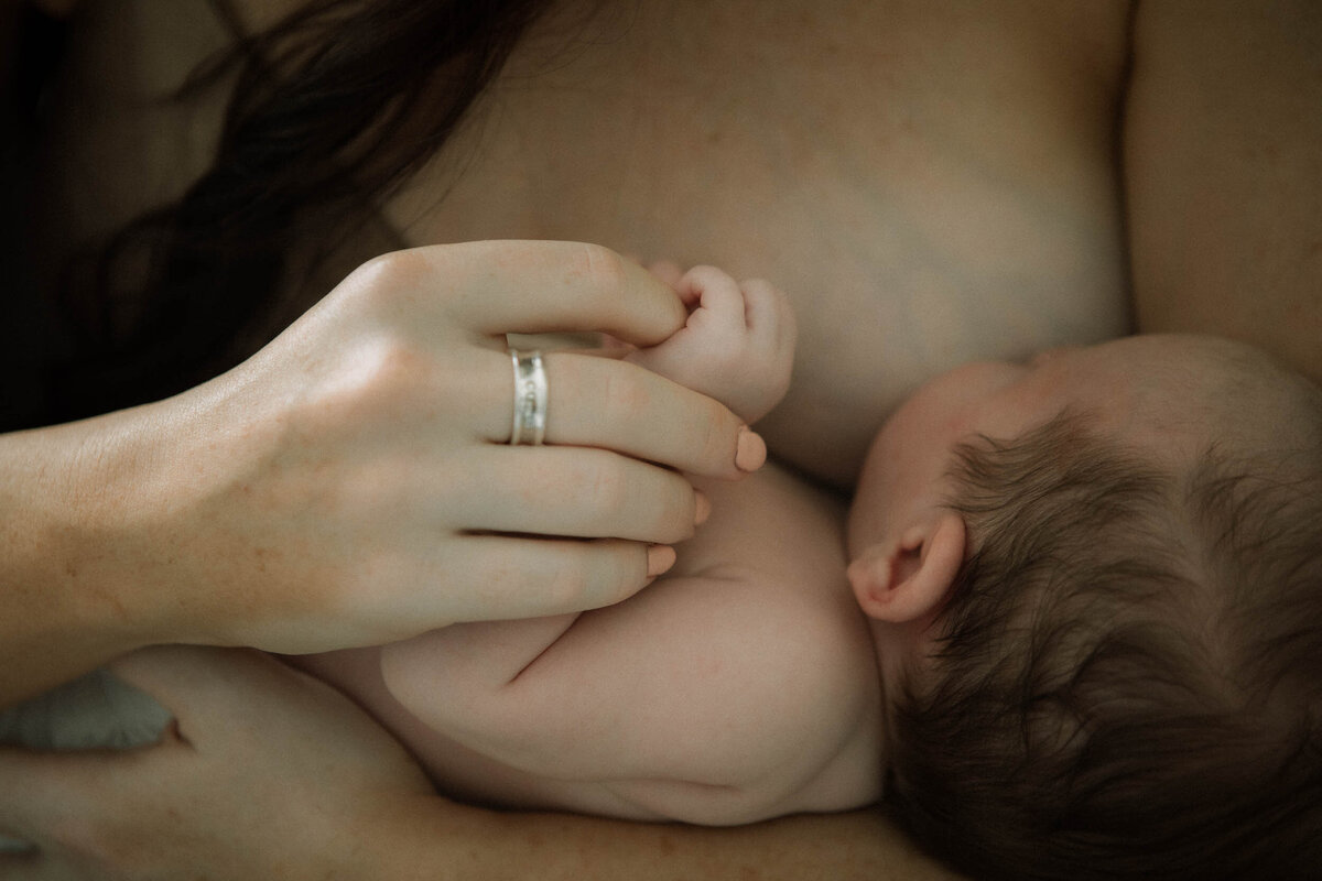 Newborn breastfeeding detail with mother’s hand wearing a wedding ring resting protectively on baby’s head.