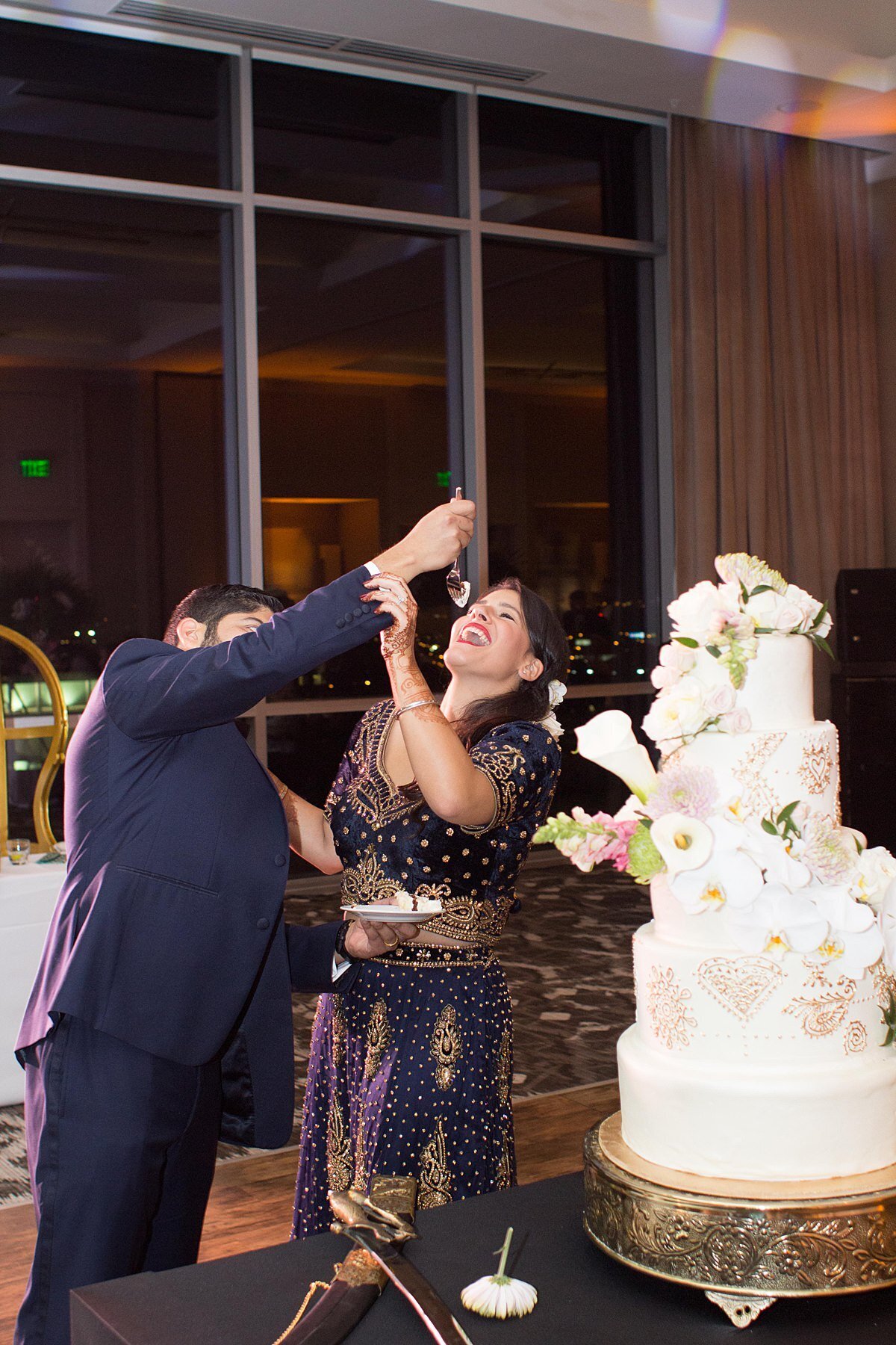 Indian Bride wearing a navy saree with gold detail and Hindu groom wearing a navy suit eat wedding cake. The Indian Wedding cake is white fondant with gold mandala detail and white orchids sitting on a gold cake stand.