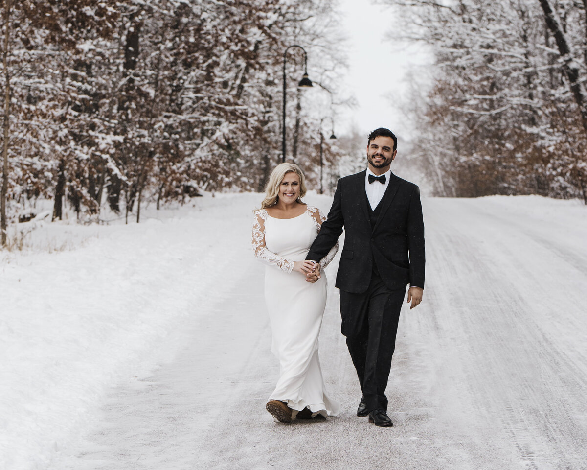 A couple dressed in wedding attire walking hand in hand on a snowy path, with trees covered in snow and a street lamp by the side taken by jen Jarmuzek photography a Minneapolis wedding photographer
