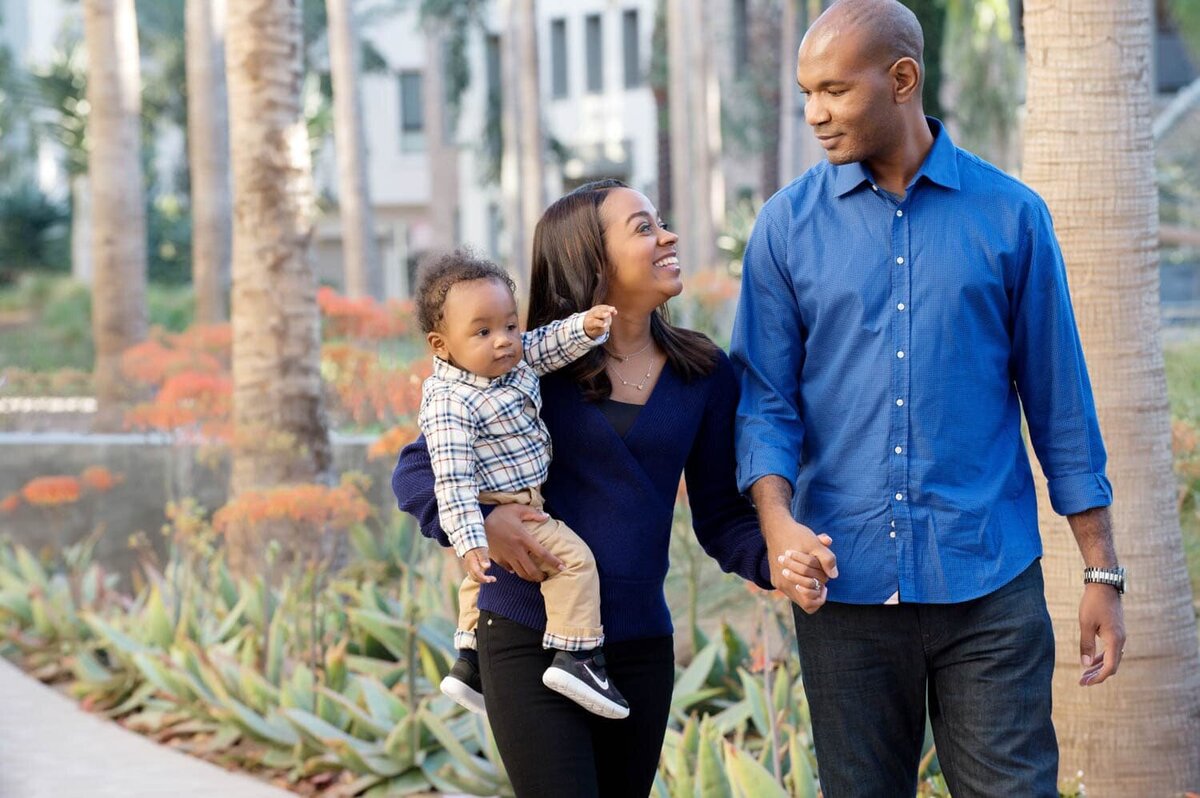 Father and Mother walking near palm trees while holding hands and their young son.