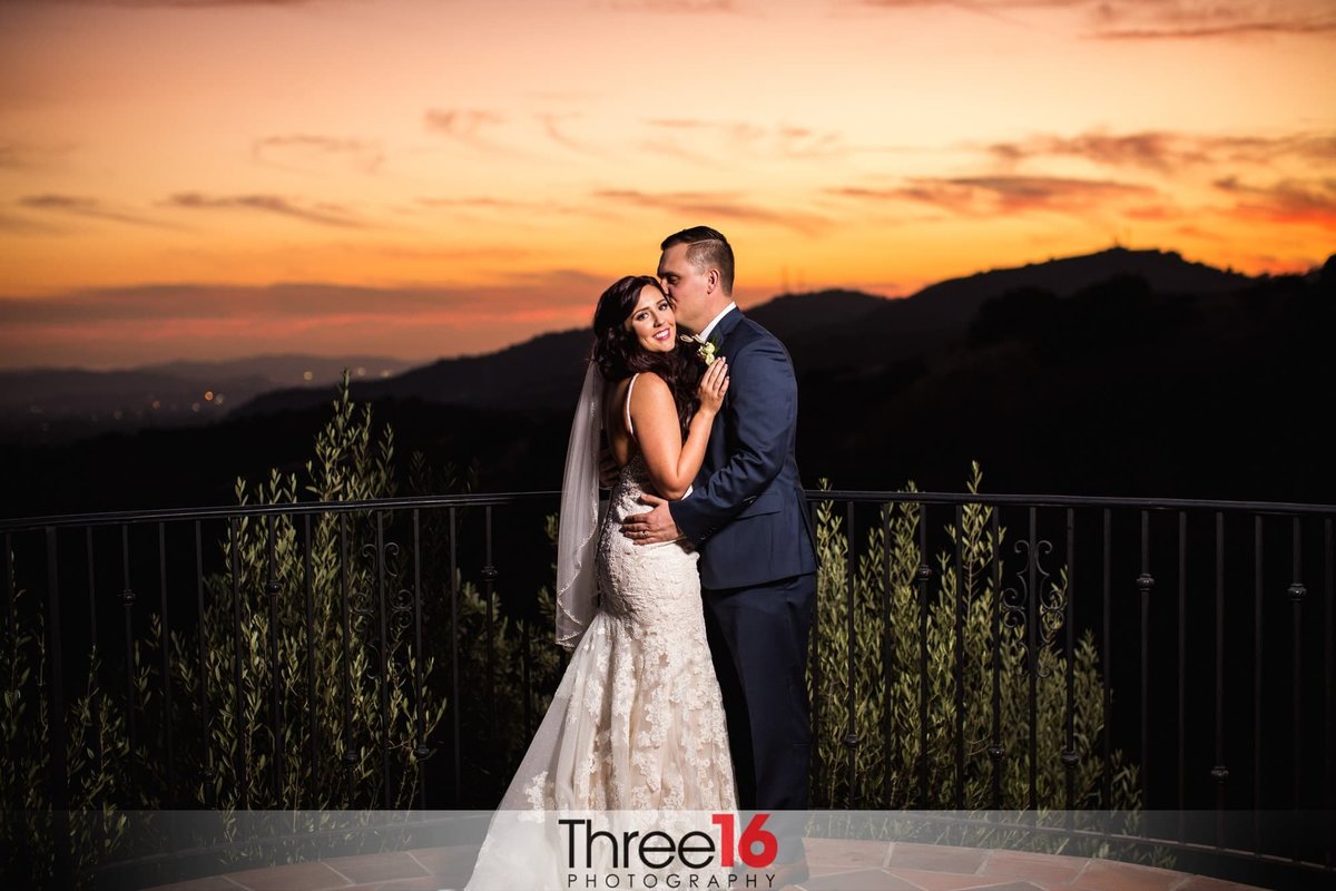 Groom kisses his Bride's cheek during photo shoot overlooking the hills with the sun setting