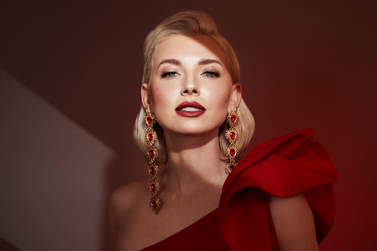 Glamorous portrait of Melissa Goodwin in a bold red outfit with statement earrings, captured by Ylva Erevall under dramatic lighting.
