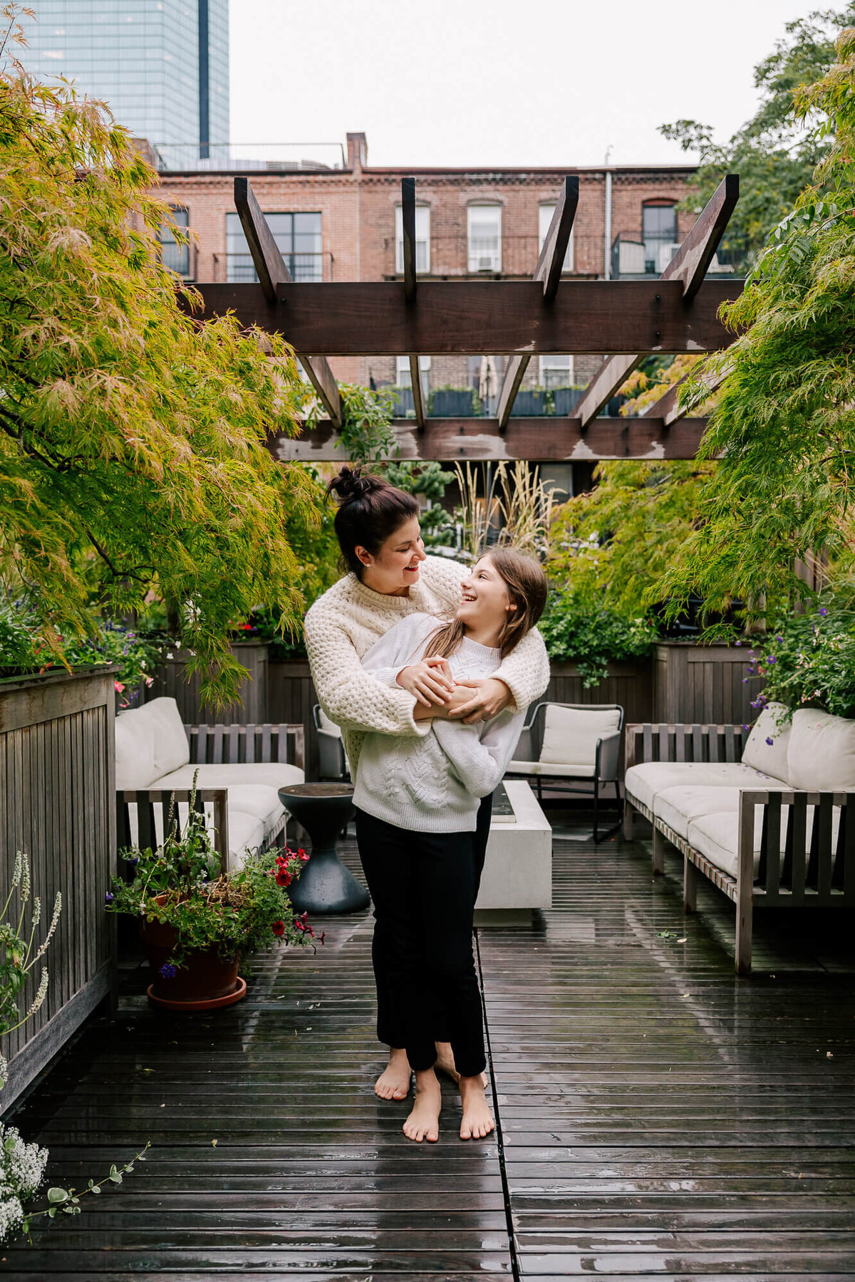 Dark-haired mother holds her pre-teen daughter from behind, as they stand on their rooftop deck surrounded by plants. The daughter has long light brown hair and is smiling up at her mother.