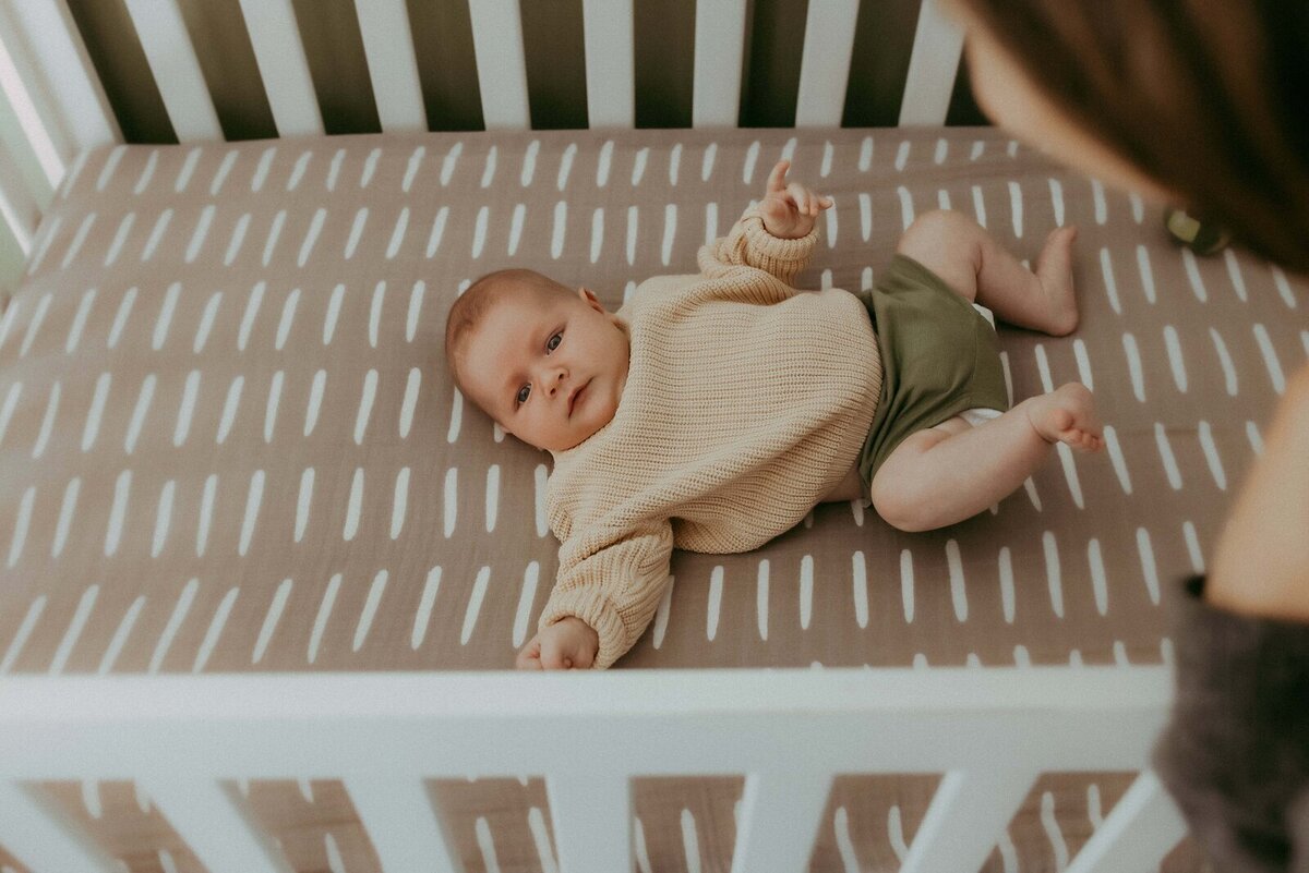 Baby lying on back in a crib with a patterned sheet, wearing a beige sweater and green shorts, looking upward.