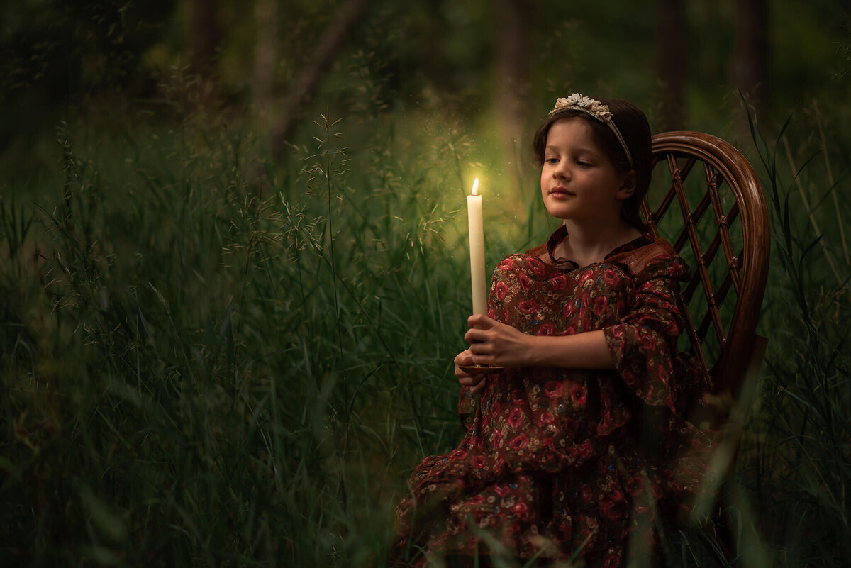 Brittany Danielle captures a portrait of a little girl sitting on a wooden chair with her knees up, holding a candle and looking at it.