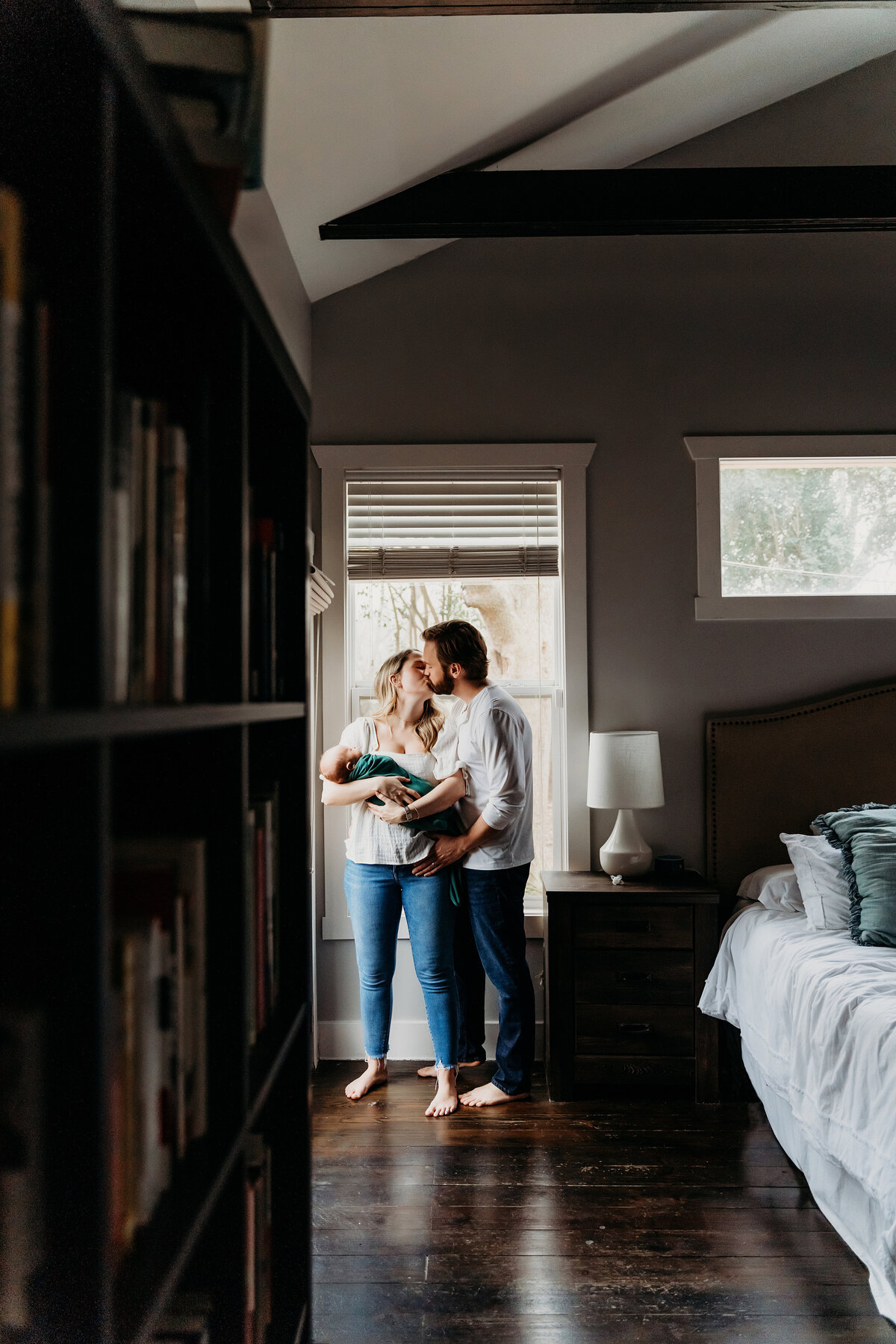 Newborn Photographer, Mom and dad kiss at the edge of the bedroom, mom holding baby