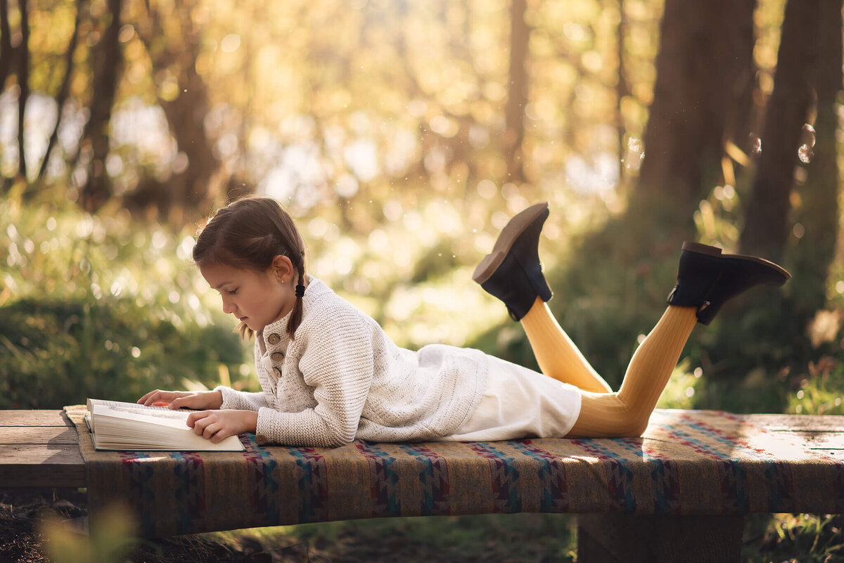 A young girl lays on her belly on a small wooden bridge in the forest reading a book with sunrays in the background.  Captured by Brittany Danielle Photography