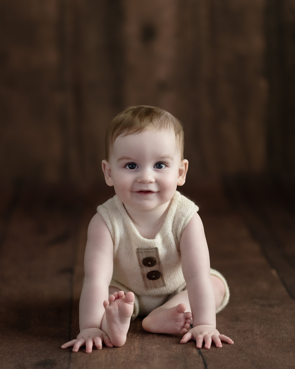 Milestone sitter photo of baby boy in a simple romper on warm wood—timeless textures, tender gaze.