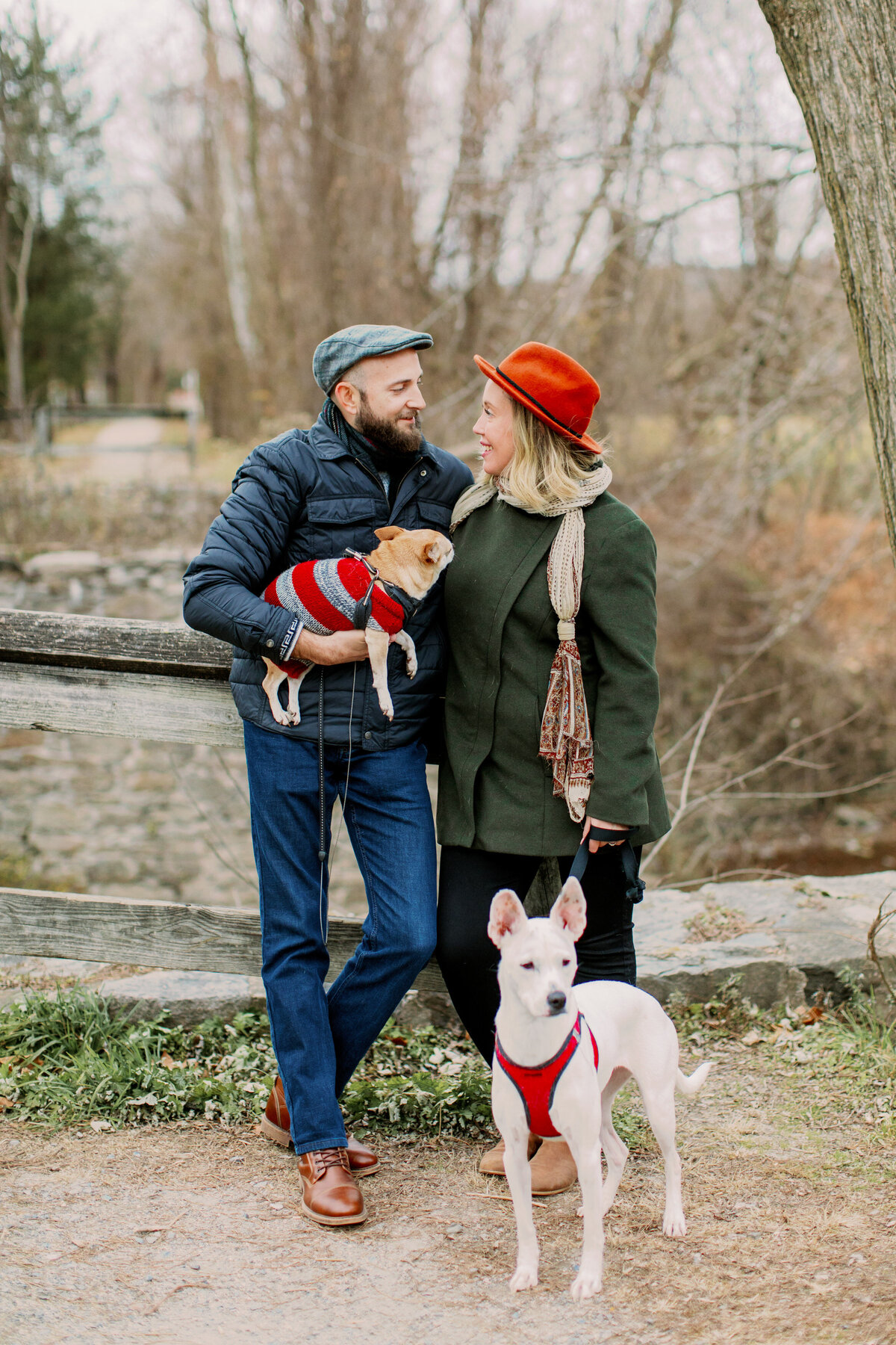 A couple with their arms around each other as one is holding a dog in their arms and the other is holding the leash of another dog 