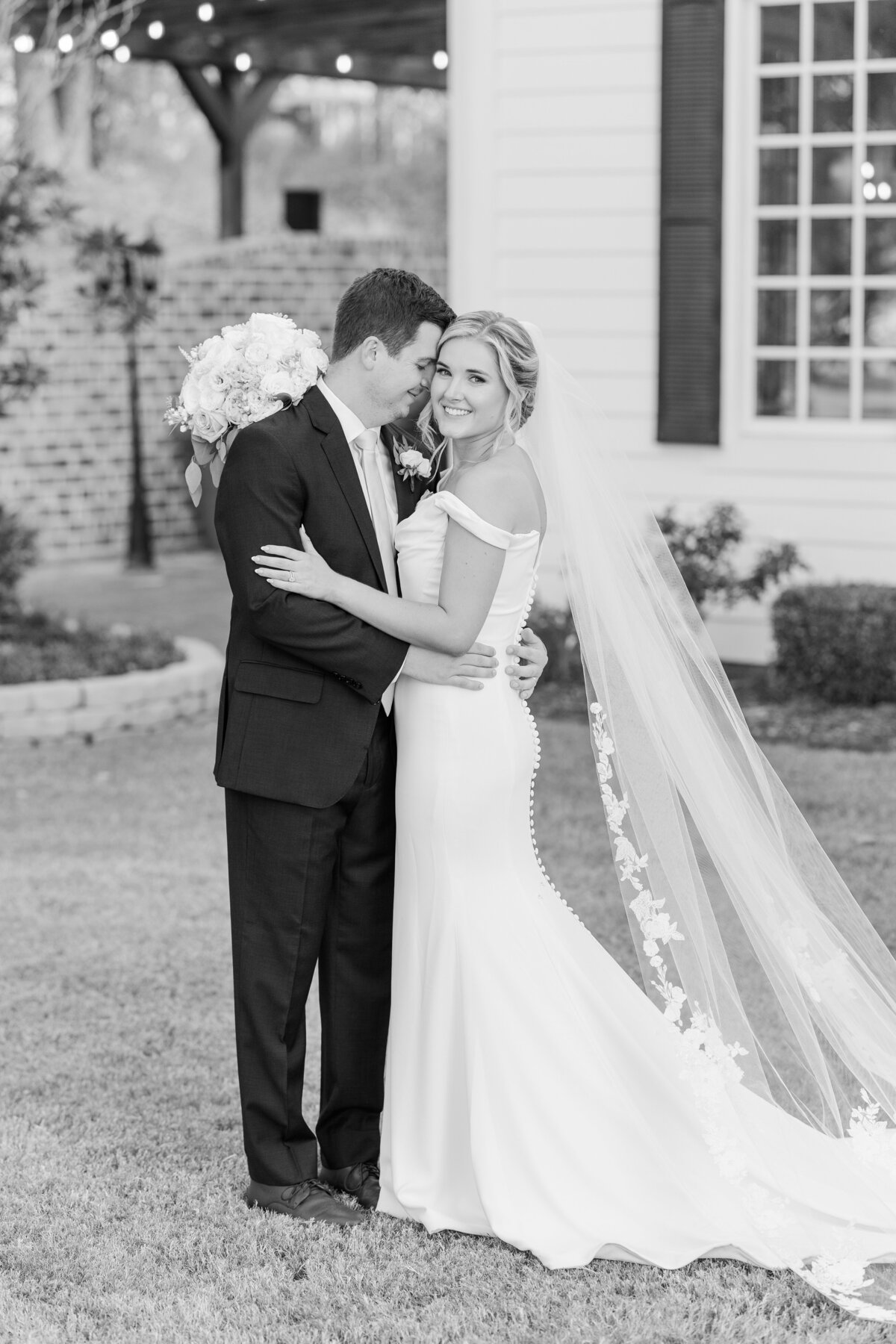 Timeless black and white wedding photo of a bride and groom sharing a romantic moment outdoors near Flower Mound, Texas. The bride’s classic gown and cathedral veil are complemented by soft floral details, perfect for couples seeking a luxury wedding photographer near Plano or Coppell.