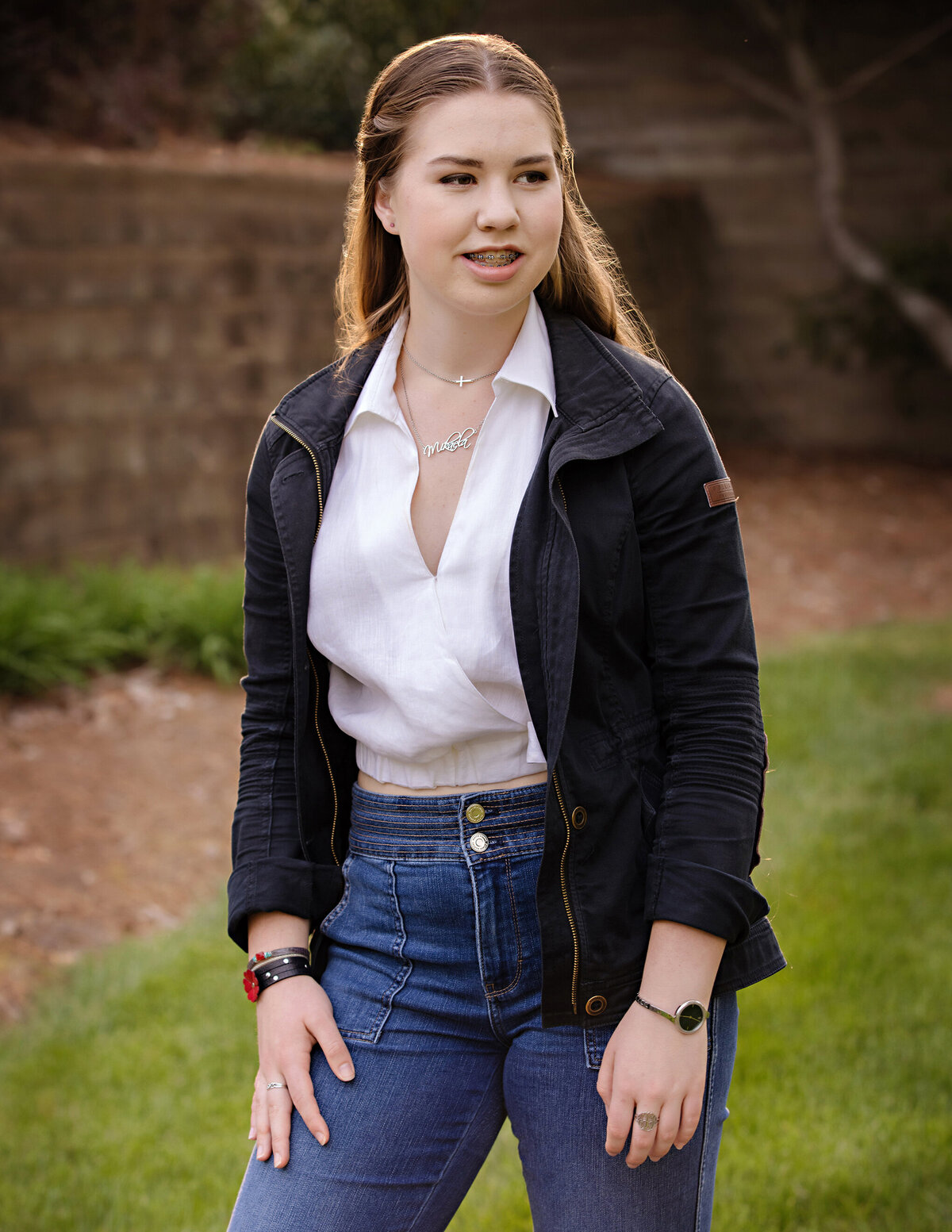 High school senior in a classic bomber jacket, white top, and jeans gazing into the distance with warm backlighting during an autumn portrait session in South Charlotte NC, captured in soft golden hour light.