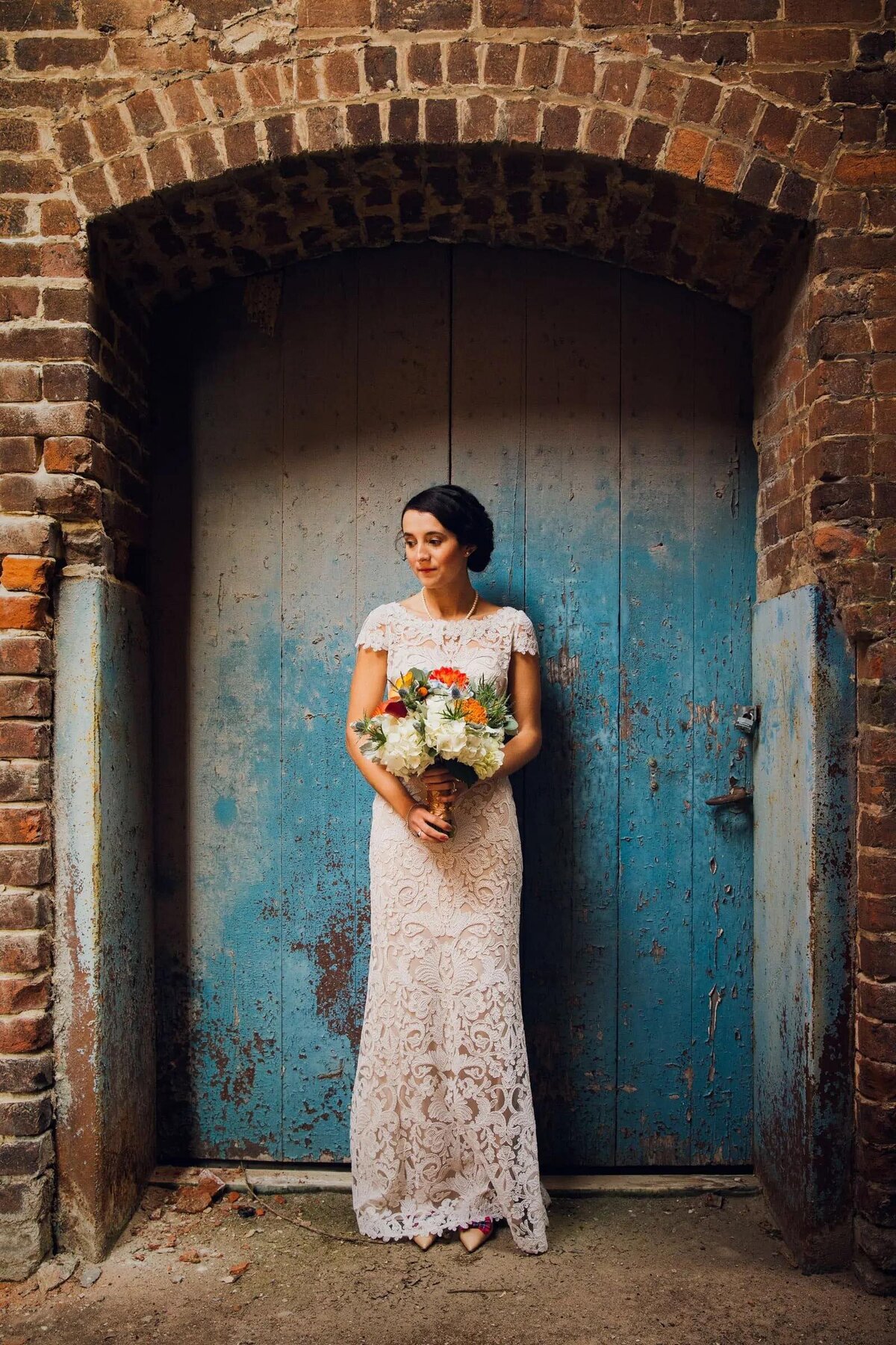 A bride stands serenely against a textured blue door, her lace gown and bouquet in contrast with the rustic backdrop.