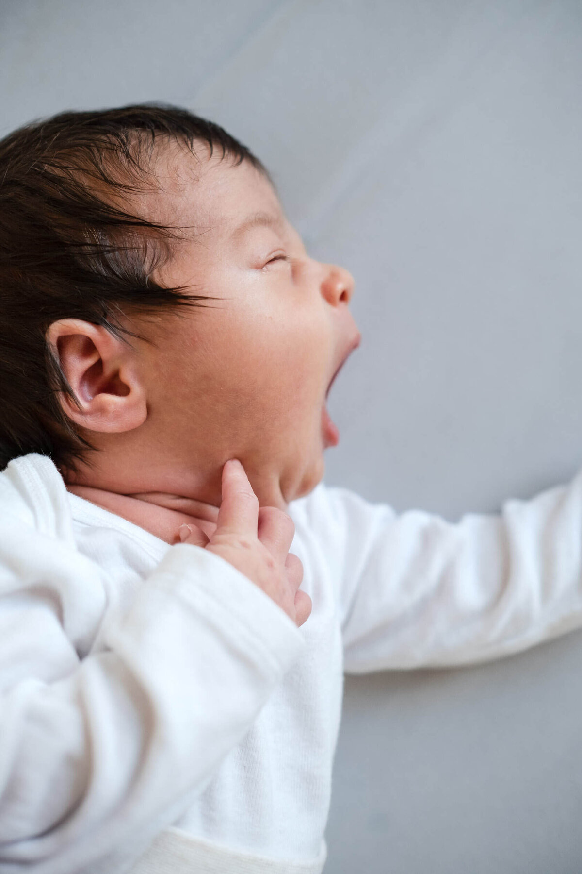 Newborn baby mid-yawn while lying on a flat surface, wearing a white onesie.
