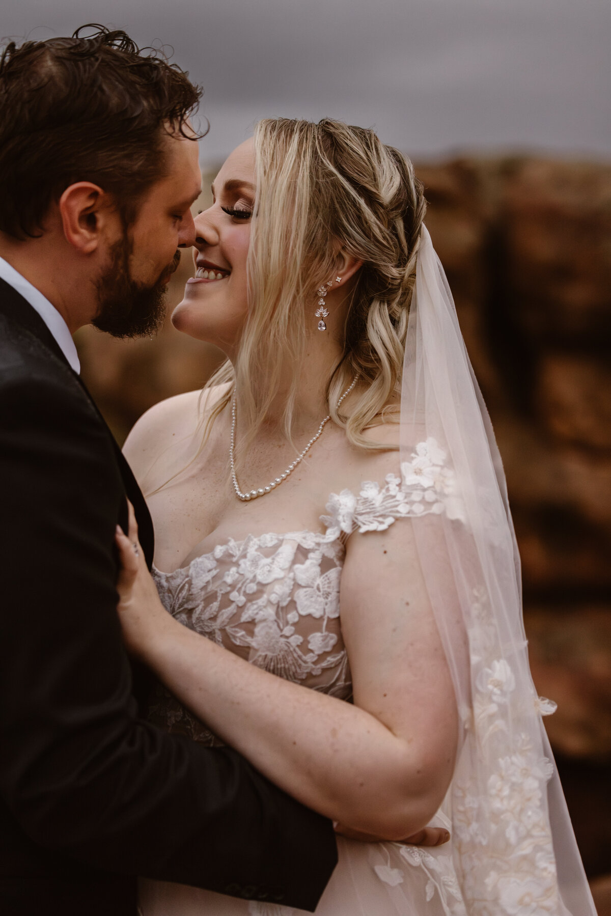 Bride and groom share an intimate moment smiling and nearly touching noses in front of red rock formations, with the bride wearing a floral lace dress and veil in Colorado.