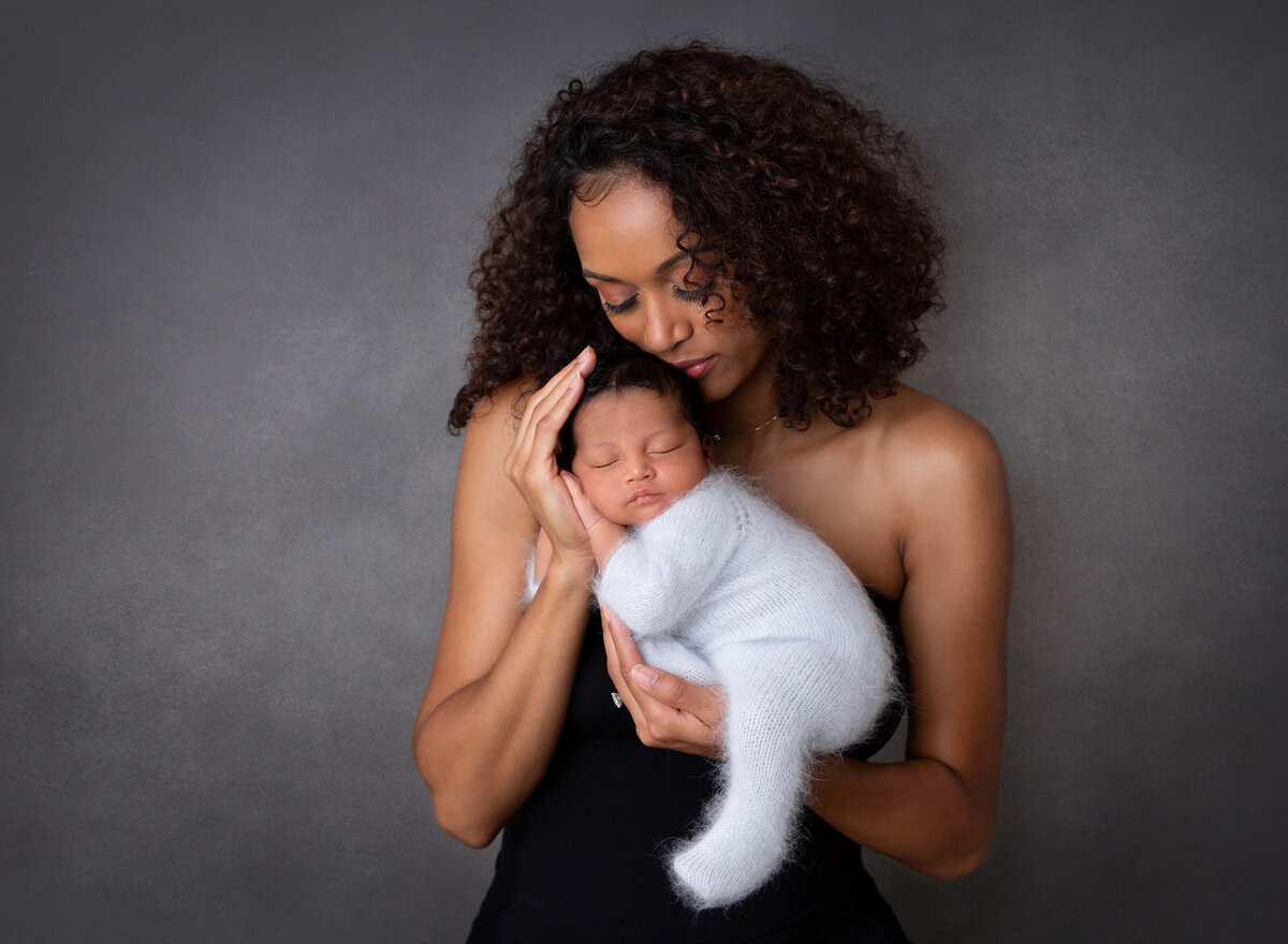 Top Brooklyn NY Newborn Photographer captures new mom holding baby in studio newborn session. Baby is wearing a white knit onesie and is lying on his belly in mom's arms. Baby's had is atop of mom's hand and he is resting his cheek on his hand. Mom has her cheek against baby's head and eyes are closed.
