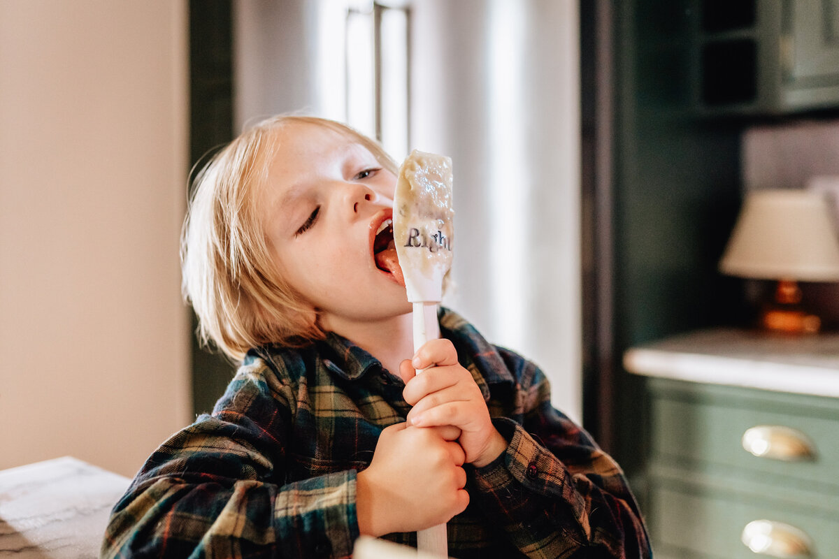 A young child in a plaid shirt licking batter from a spatula in a kitchen, enjoying the sweet treat with eyes closed in contentment.