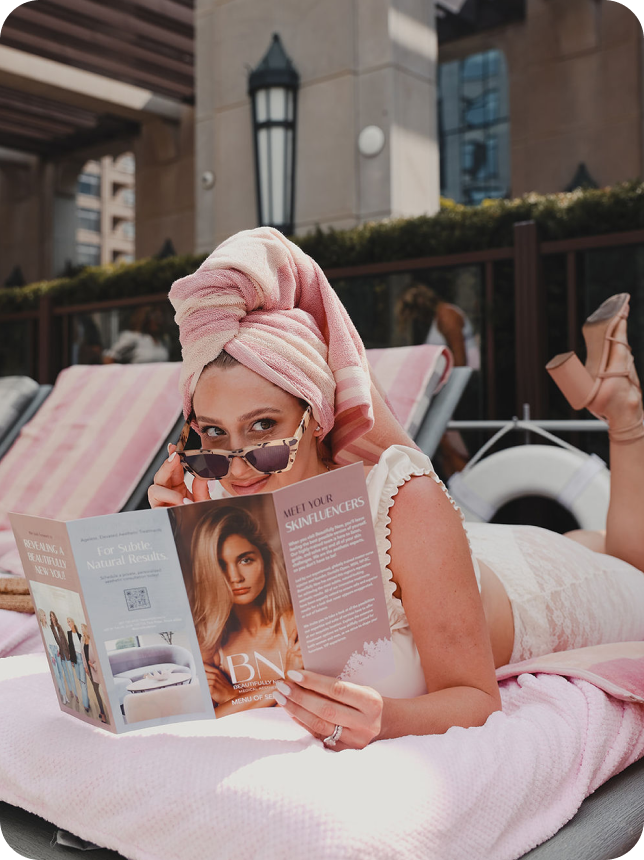 A woman in a bathing suit and a pink towel on her head reading a pamphlet for one of Mello Social's photoshoots. 