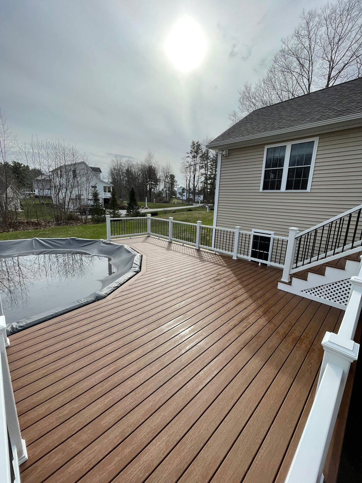 A beautiful dark brown deck with white railings and trellis stairs facing around a pool done by a Shrewsbury MA Deck Builder