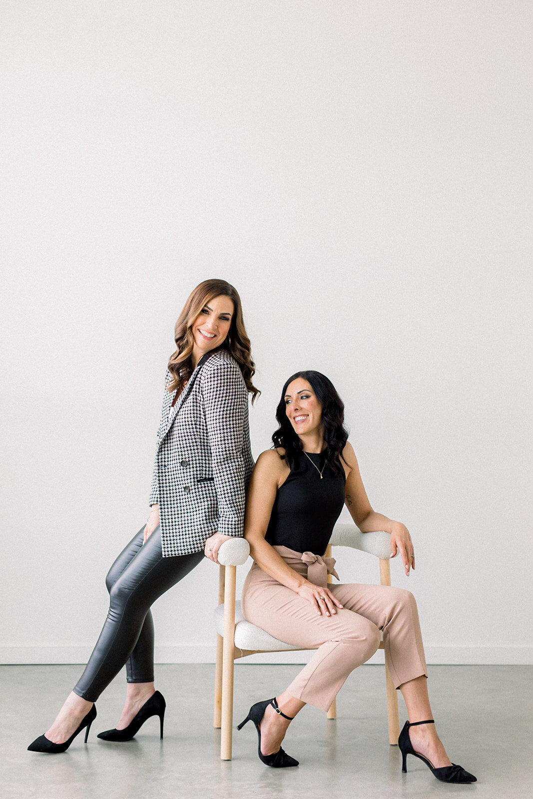 Two Calgary business women with dark hair laughing while posing on white chair