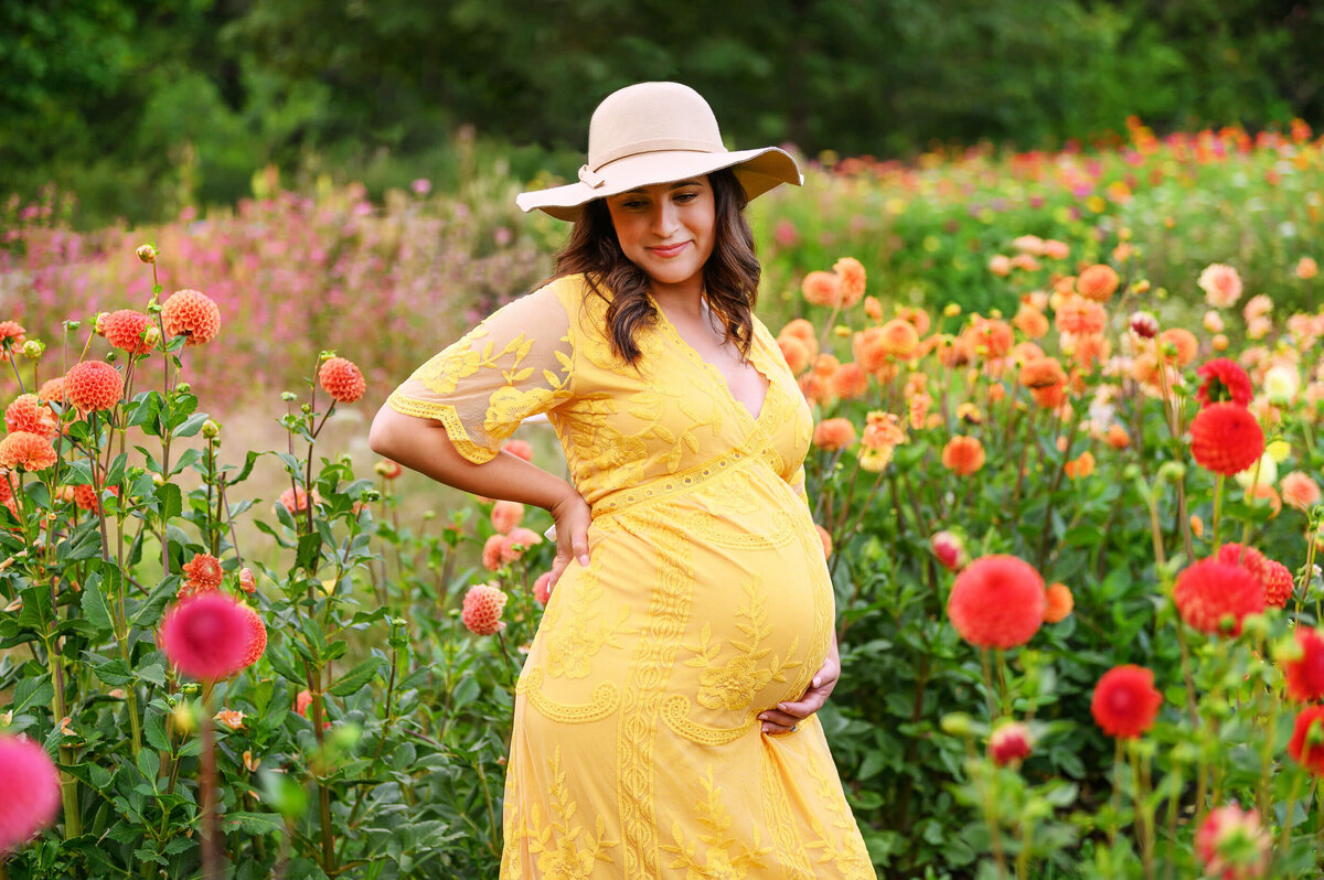 pregnant woman holding her belly wearing a yellow dress with a beige hat on in at a flower farm for her outdoor newborn photography session in Burlington, Ontario