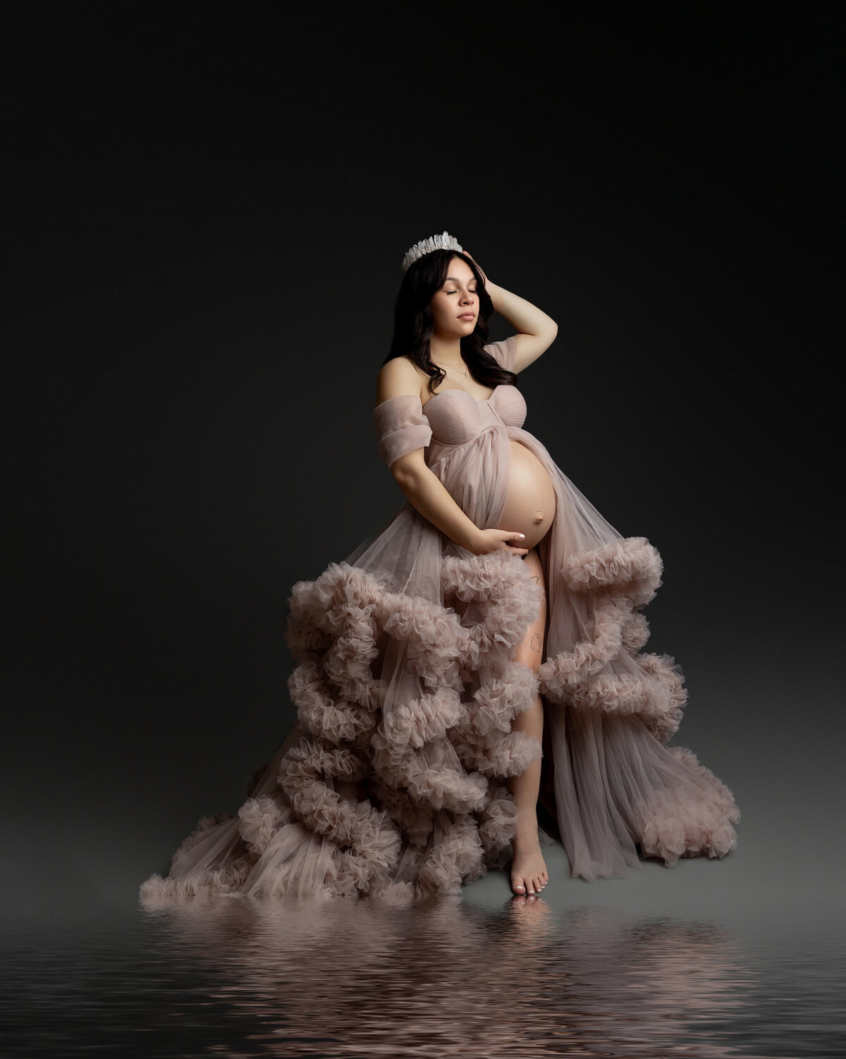 In a soft pink ruffle dress, she poses against a dark backdrop, with her reflection beautifully captured in the ground, adding depth and elegance to the moment. Captured by Inland Empire maternity photographer Kaitlyn Dawn Photography.