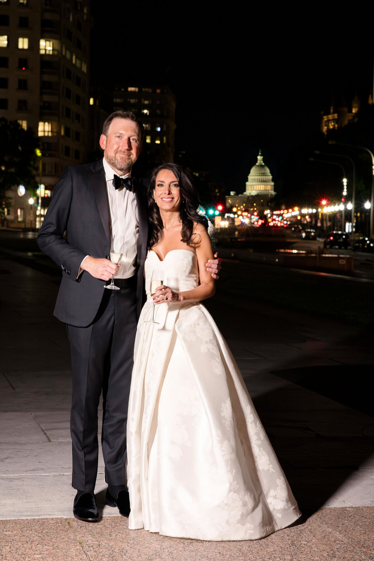A couple dressed in formal attire stands on a city street at night. The man wears a tuxedo, and the woman wears a white strapless gown. They are smiling and holding champagne glasses, with a lit domed building visible in the background.