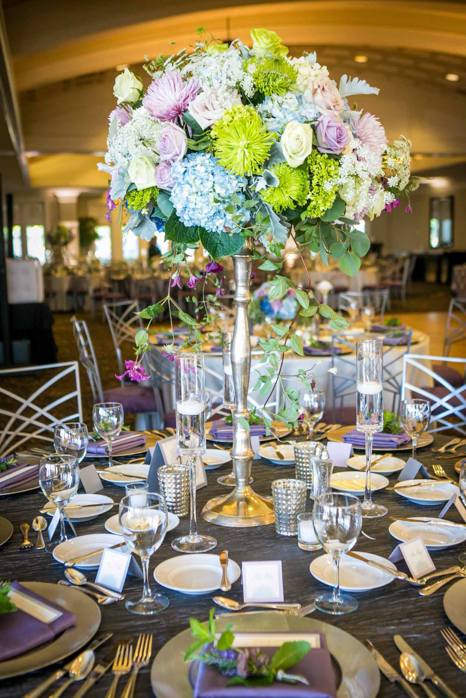 large centerpiece of blue hydrangea, purple roses, green chrysanthemums, , and trailing purple clematis, with floating glass candle holders