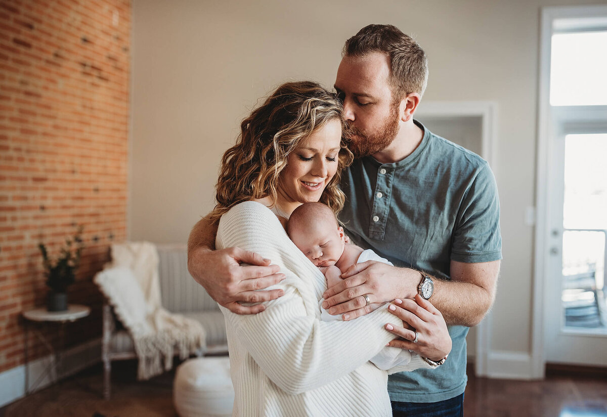 Husband hugging wife and newborn in light filled room in Baltimore Maryland