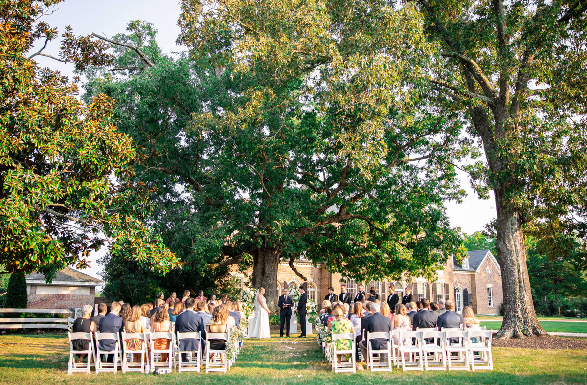 A wedding ceremony in an outdoor garden setting. Guests sit on white chairs facing a couple under a large tree. The background features a brick building surrounded by lush greenery. Bright sunlight filters through the leaves.
