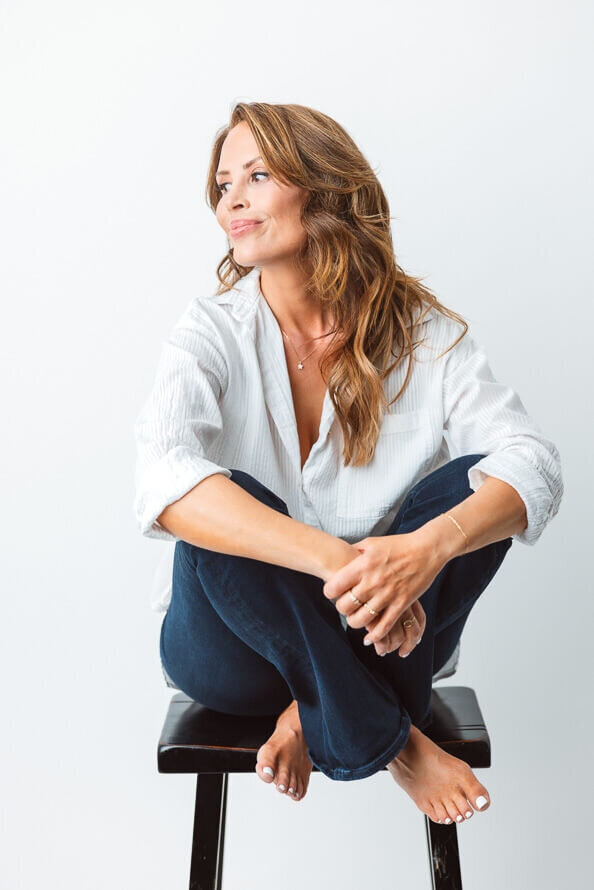 Girl in white shirt and jeans pulls legs up and looks off to side in a white photos studio.