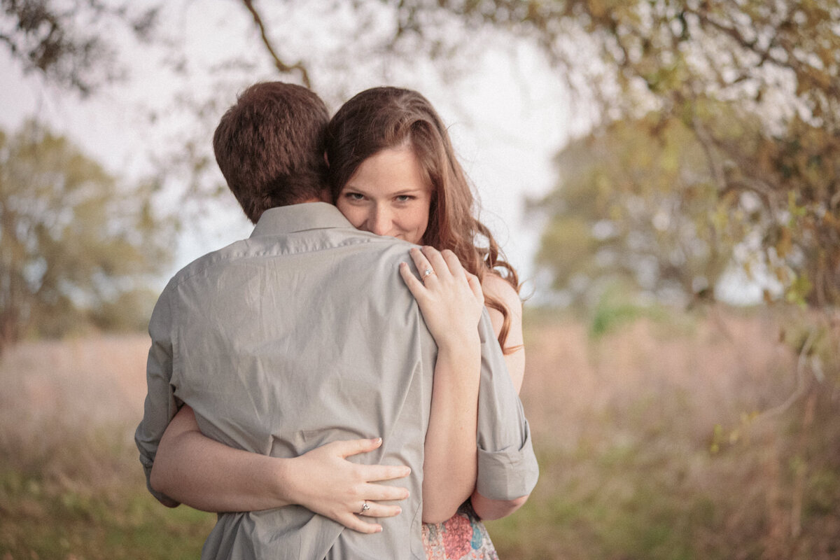 A woman embraces her fiancee, peeking over his shoulder at the camera while displaying her engagement ring