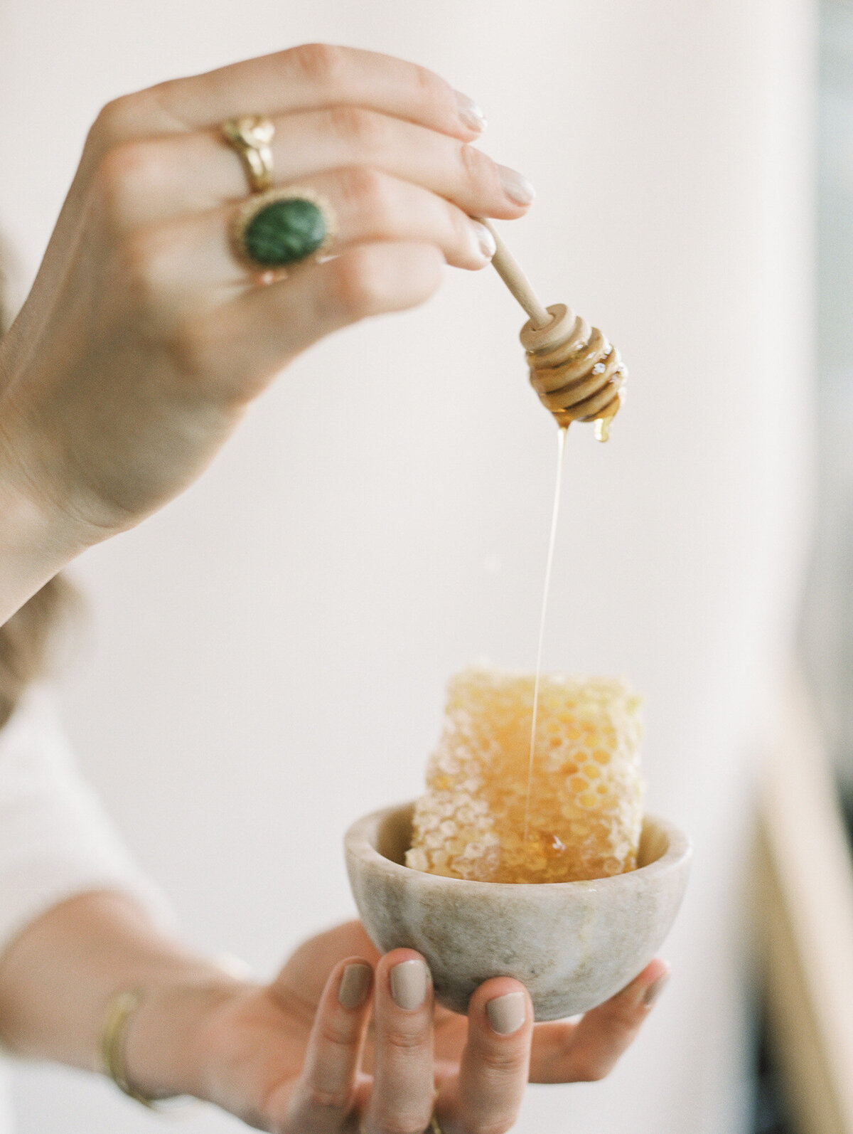 woman's hand with gold jewelry, green stone holds honey dipper with dripping honey over honeycomb in bowl, film brand photo