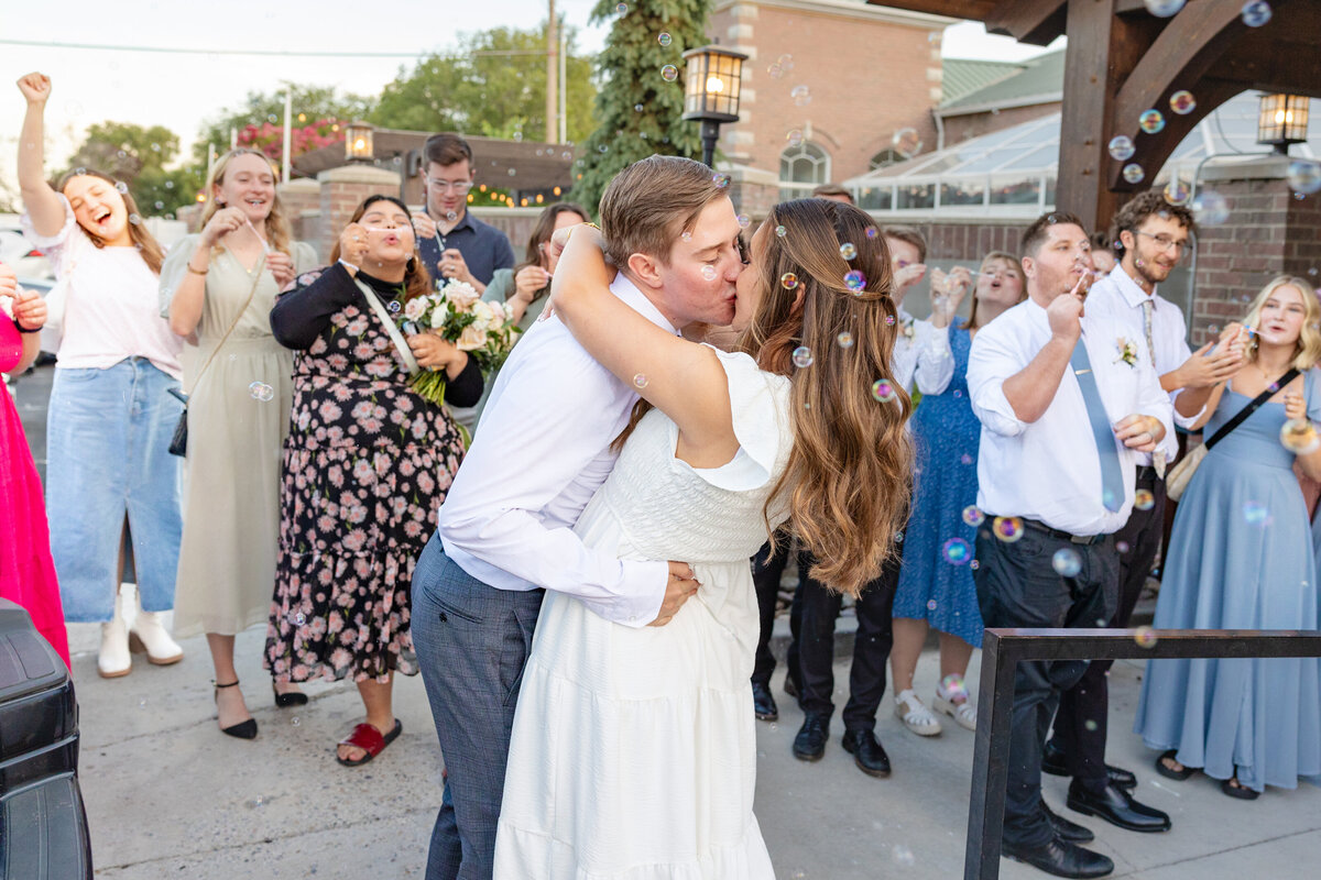 A bride and groom share a passionate kiss as guests blow bubbles around them in a joyful wedding send-off.
