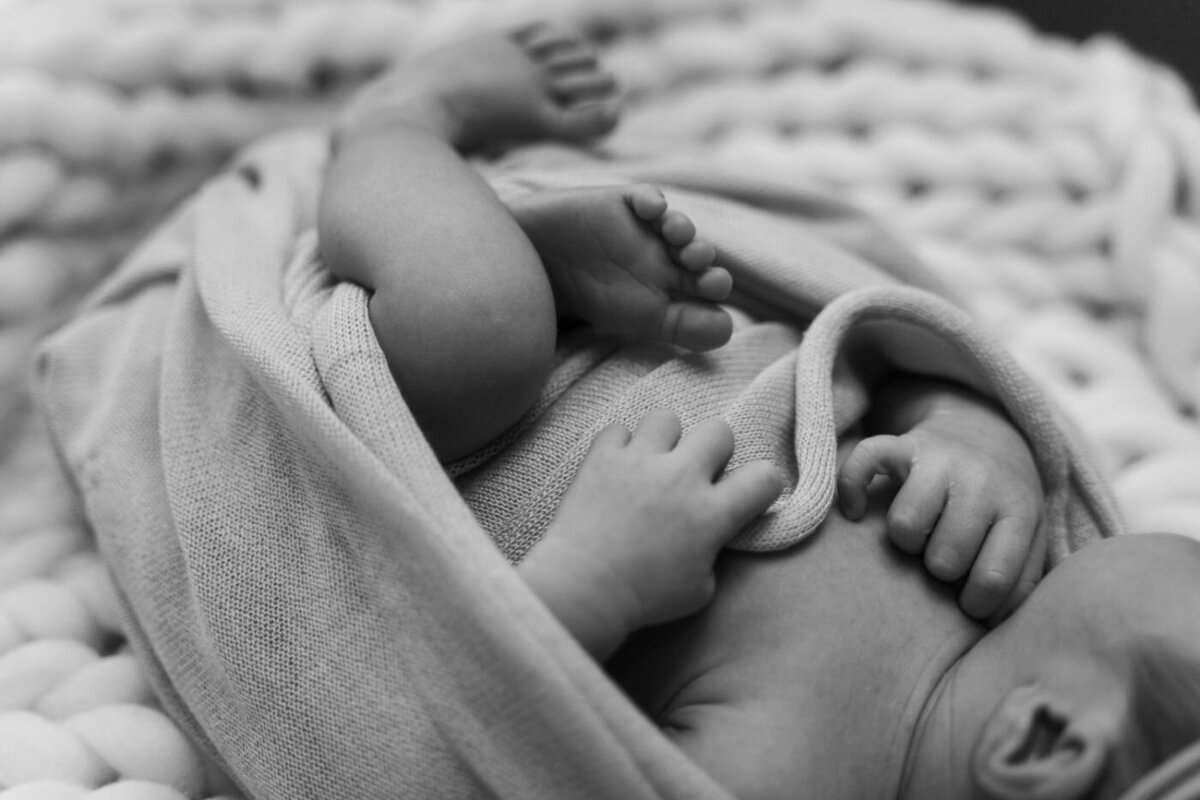 Newborn detail shot – Black and white photo of tiny newborn hands and feet peeking out from a soft knit swaddle on a chunky blanket.