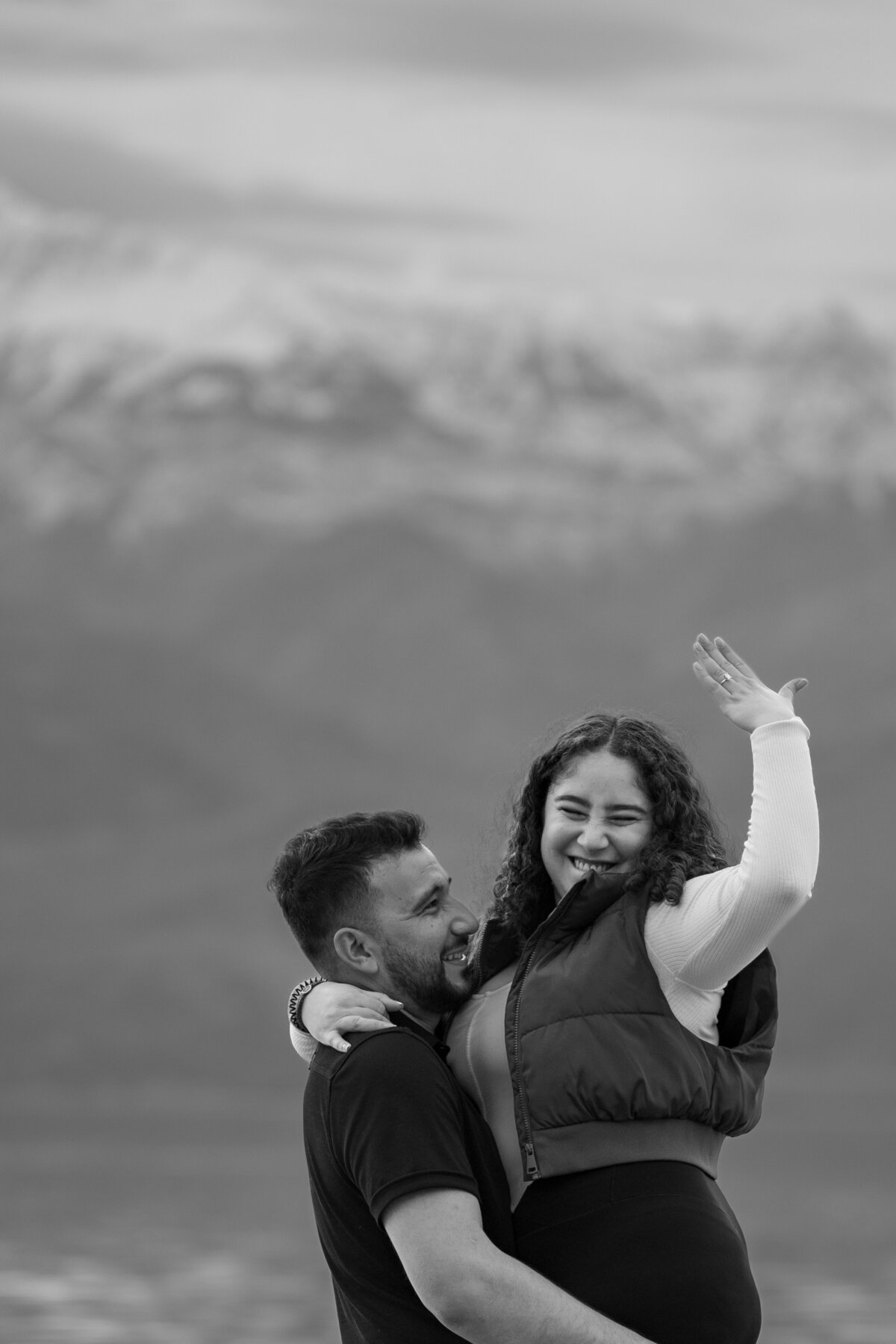 A couple hugging as one holds up their hand showing off an engagement ring