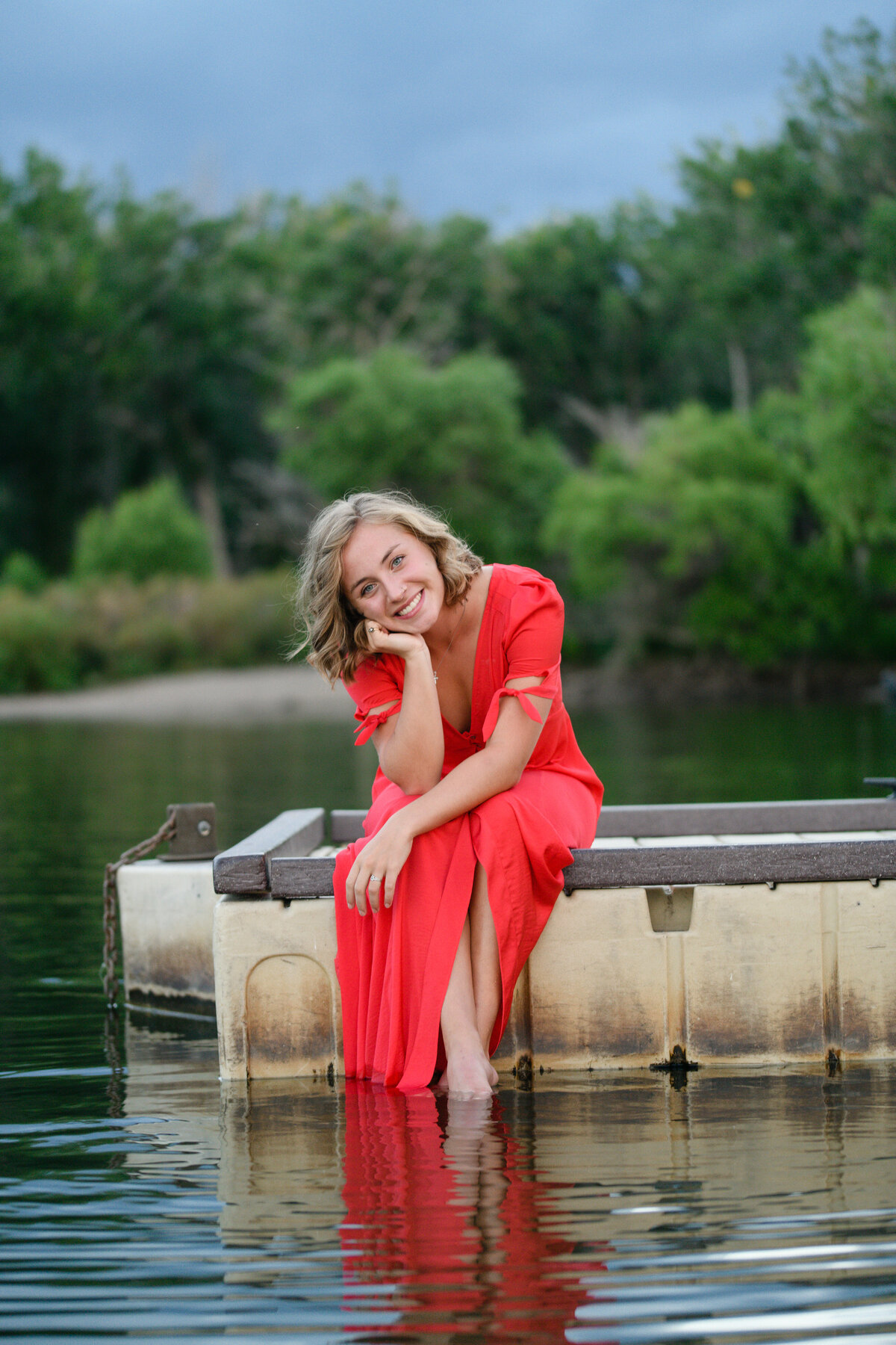 senior yearbook photos with young woman in a red dress sitting on the edge of a dock with her feet in the water of a lake  captured by denver senior photographer