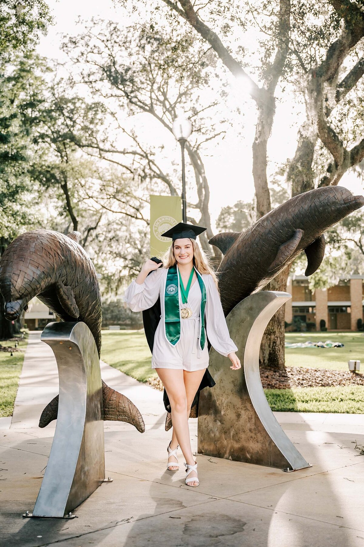 Graduate holding her gown and wearing a cap between dolphin statues at JU