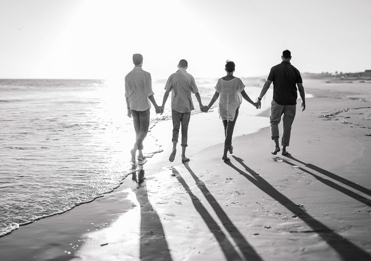 Four people holding hands walk along a beach at sunrise or sunset, casting long shadows on the sand. They face the ocean, creating an evocative scene captured by a Fayetteville NC family photographer. The black-and-white composition adds a timeless touch to their shared moment.